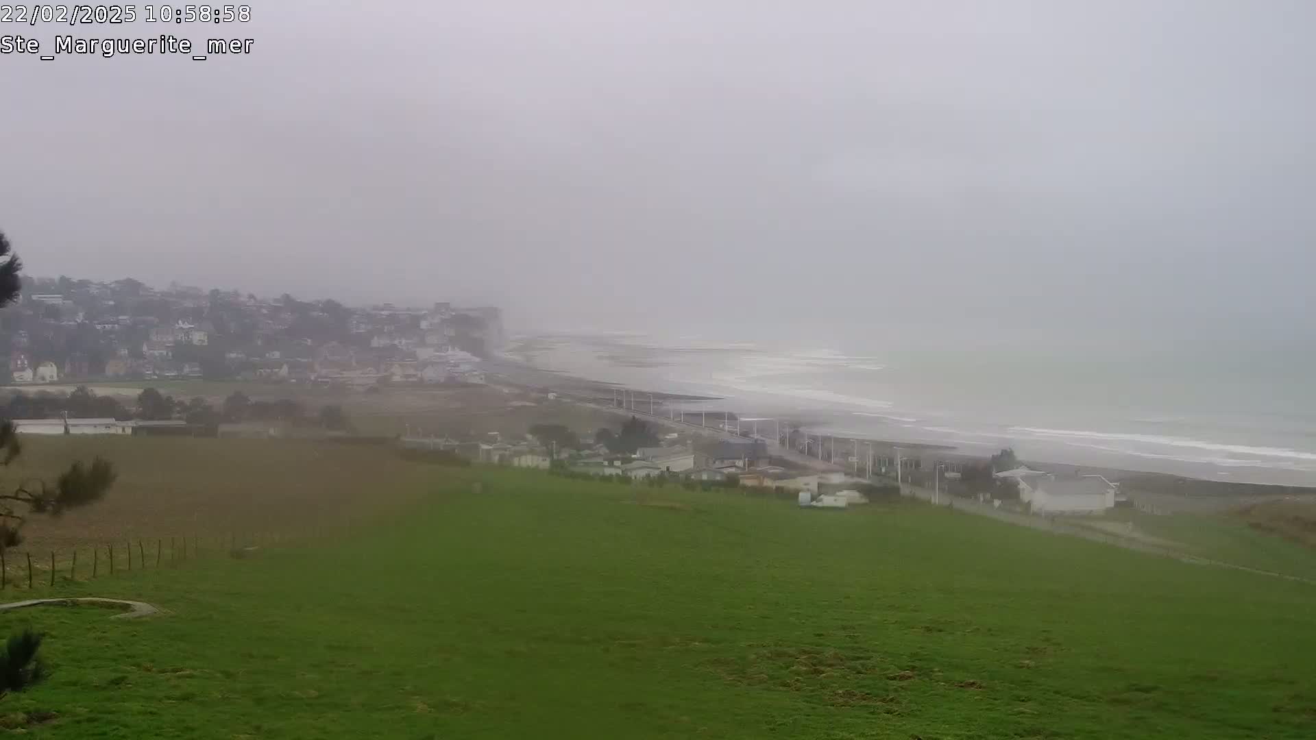 A hazy, overcast view shows a coastal town, a beach with waves, and a grassy field in the foreground.