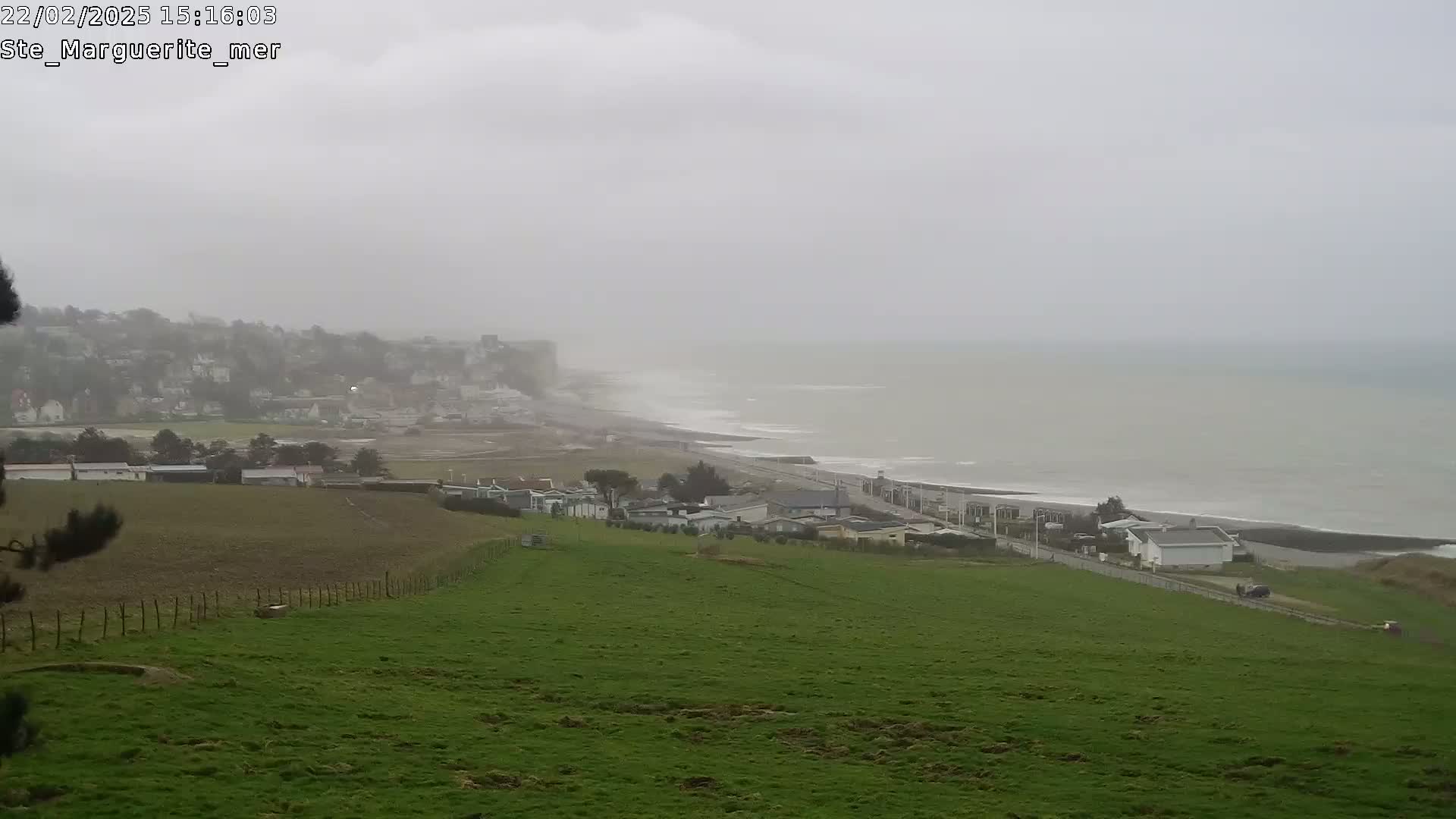 A foggy coastal scene shows a green field in the foreground, a road leading to a town near a grayish-colored sea, with buildings visible on a hill in the distance.