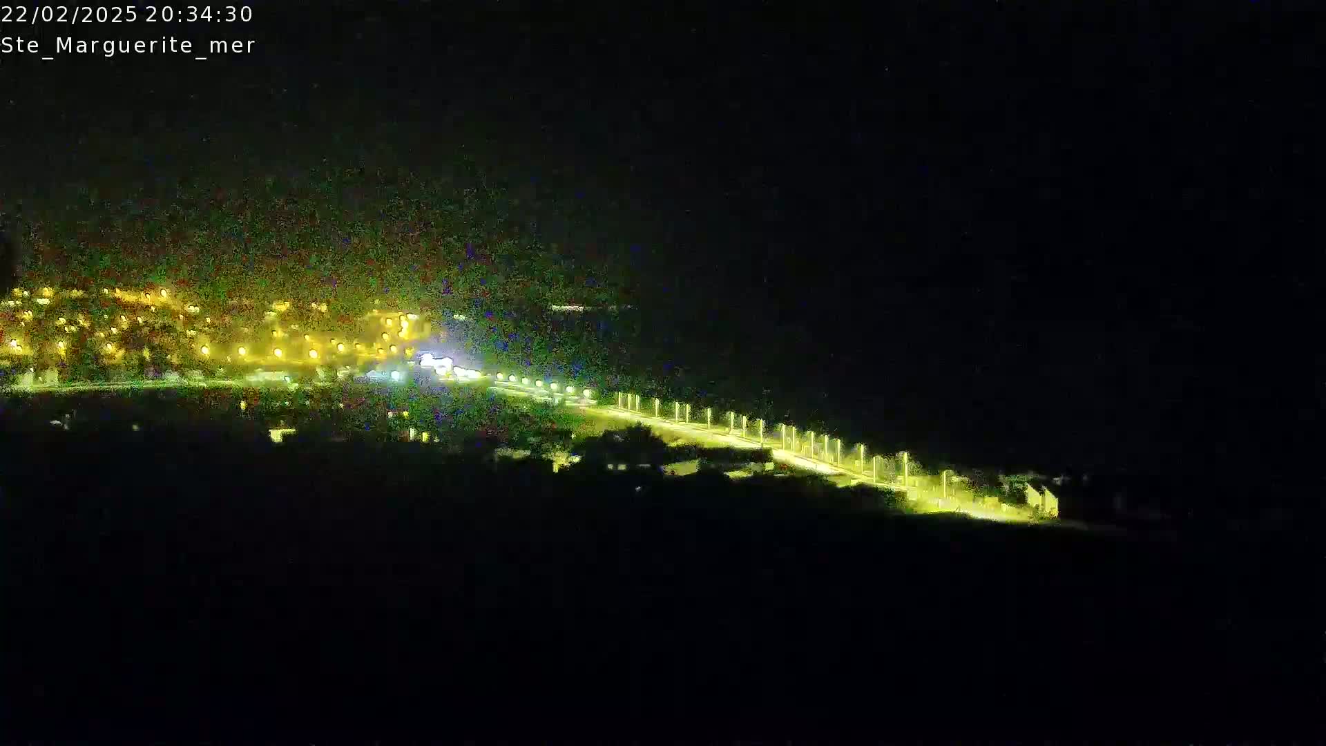 A nighttime, long-range view shows a brightly lit road winding down toward a cluster of lights in the distance.