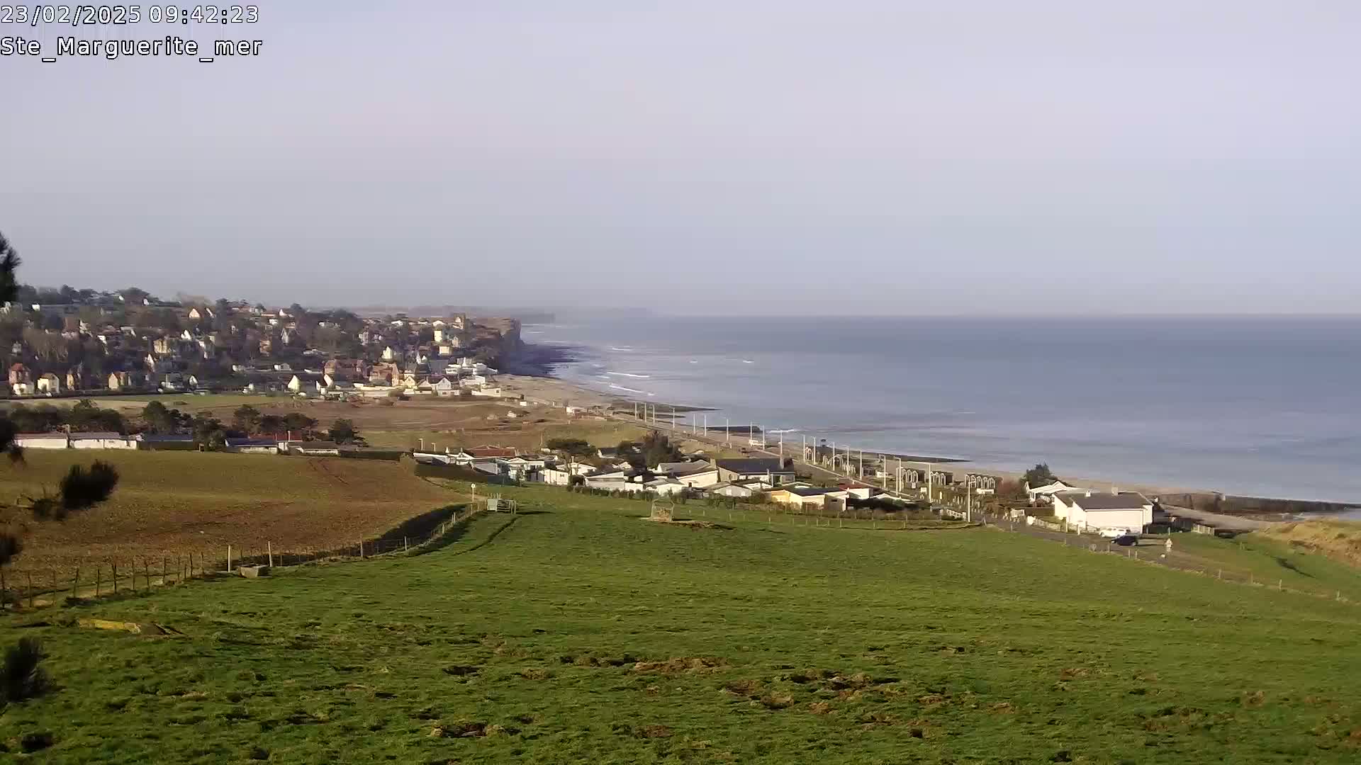 A coastal town sits atop a cliff overlooking a calm ocean, with green fields stretching between the viewer and the town on a clear day.