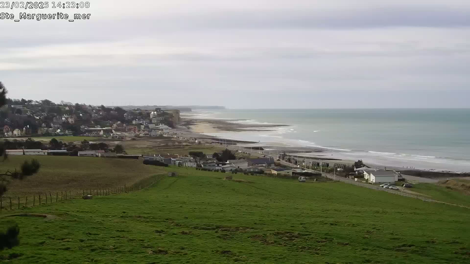 A coastal town sits atop a grassy hill overlooking a sandy beach and calm ocean under an overcast sky.