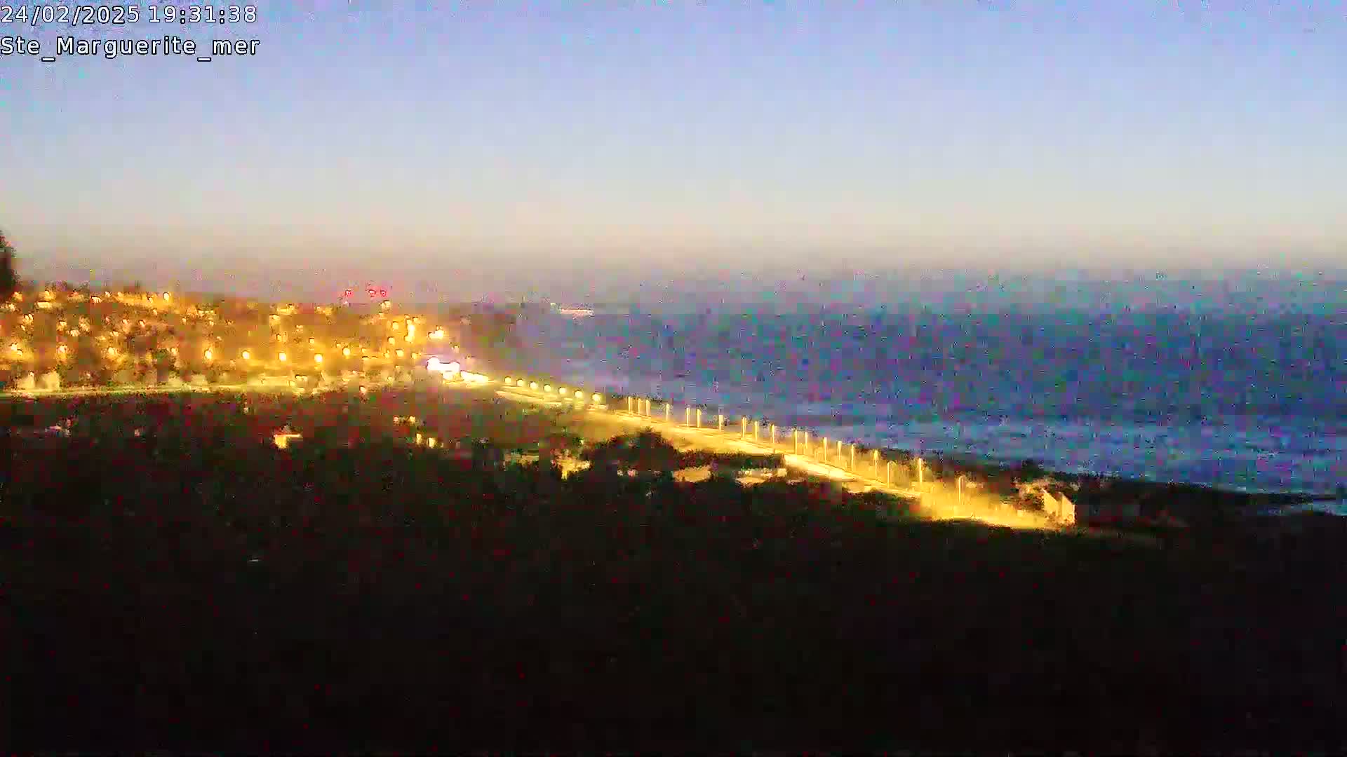 A nighttime coastal scene shows a town illuminated by lights overlooking a dark, wavy ocean under a twilight sky.