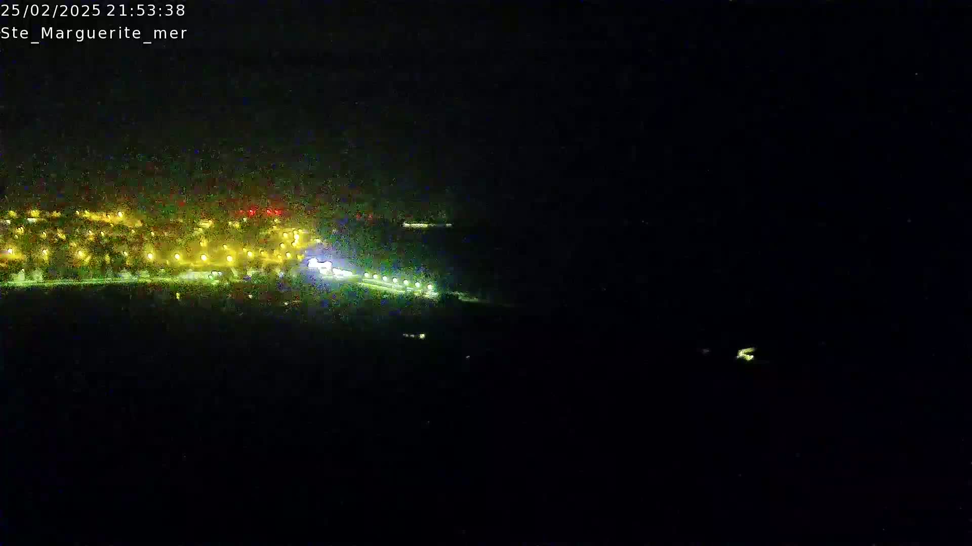 At night, a brightly lit coastal area is seen from a distance, with dark water extending into the blackness.