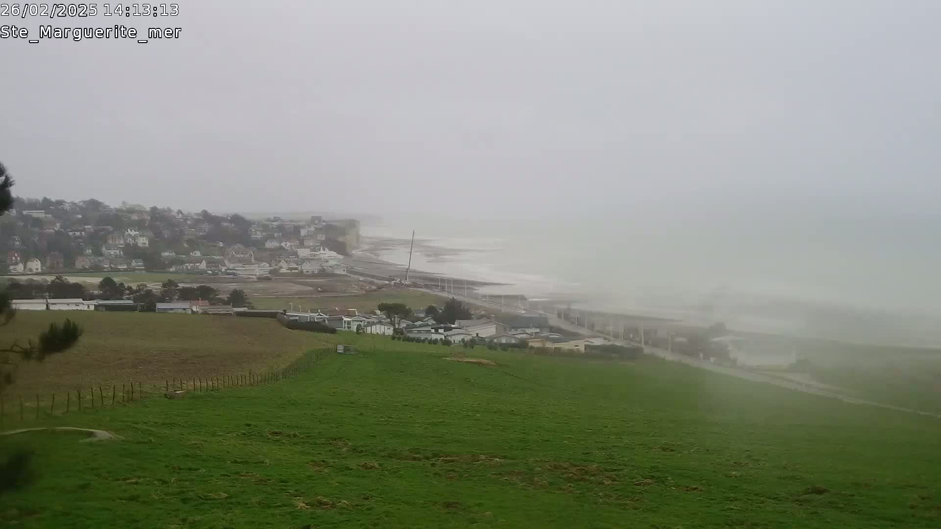 A foggy coastal scene shows a green field sloping down towards a town, which sits on a cliff overlooking a gray, wavy sea.