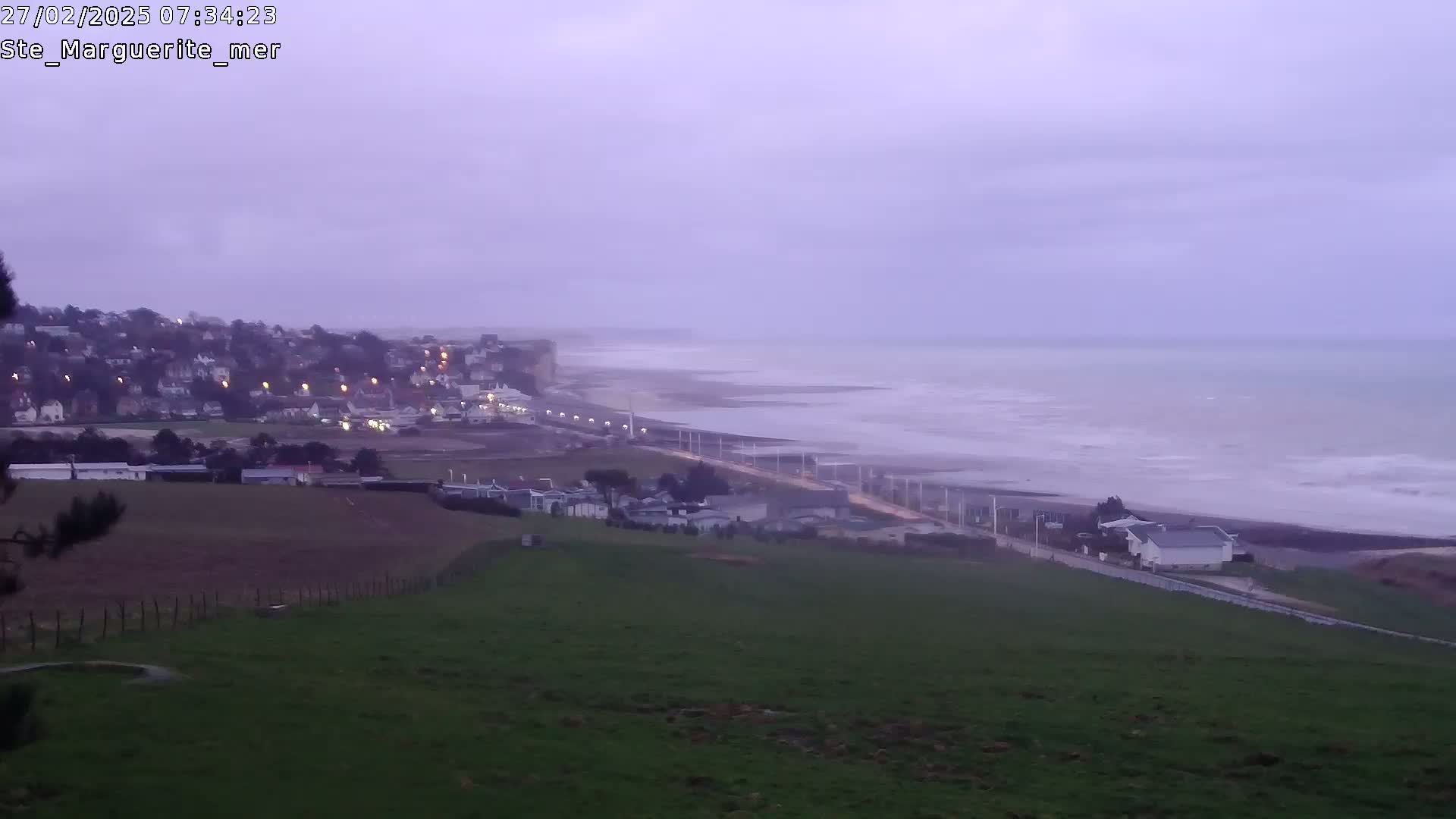 A coastal town with lights visible at twilight overlooks a beach with a choppy sea under an overcast sky.