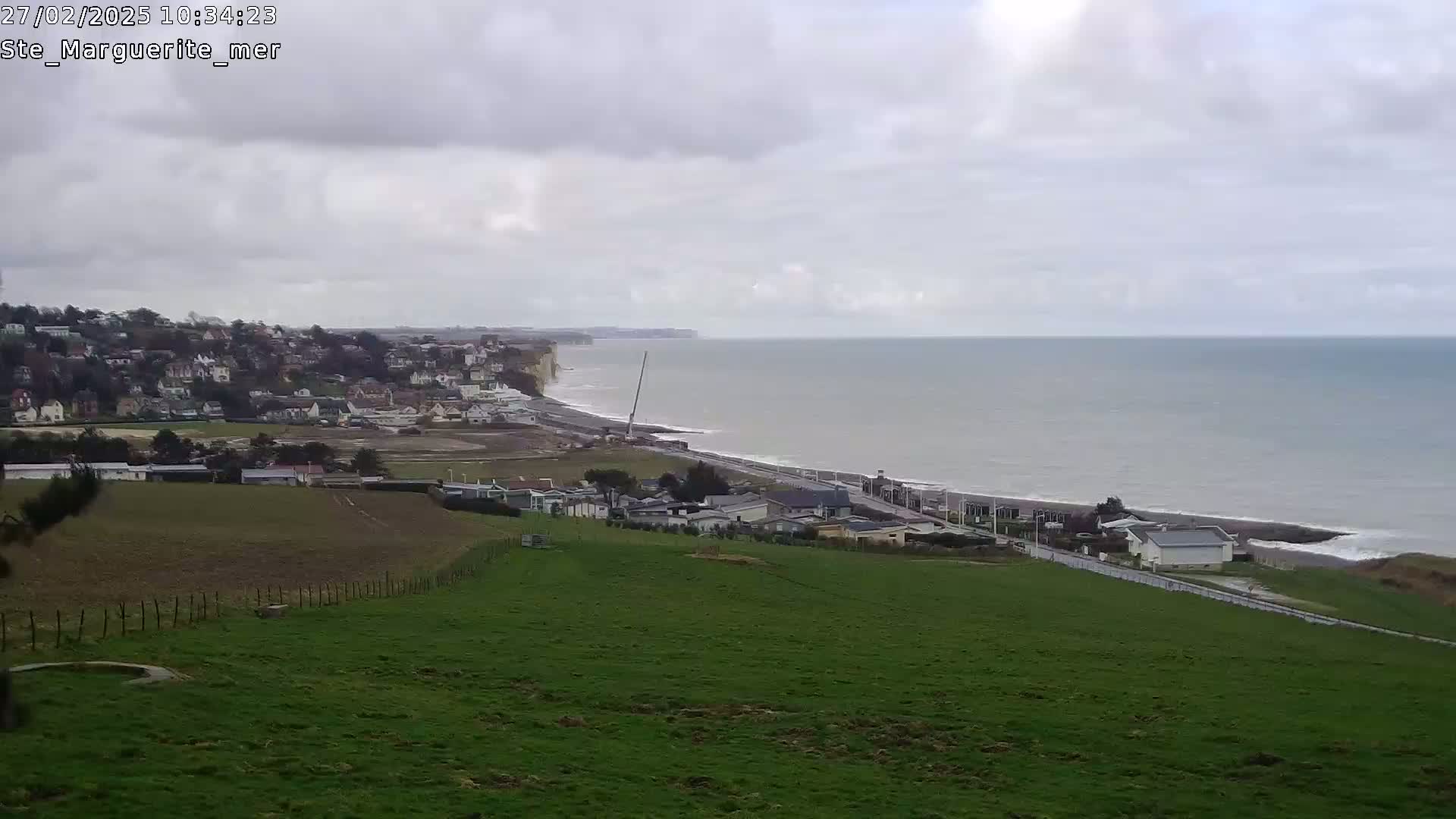 A coastal scene on an overcast day shows a green field sloping down to a town and beach, with white cliffs and a calm sea extending to the horizon.