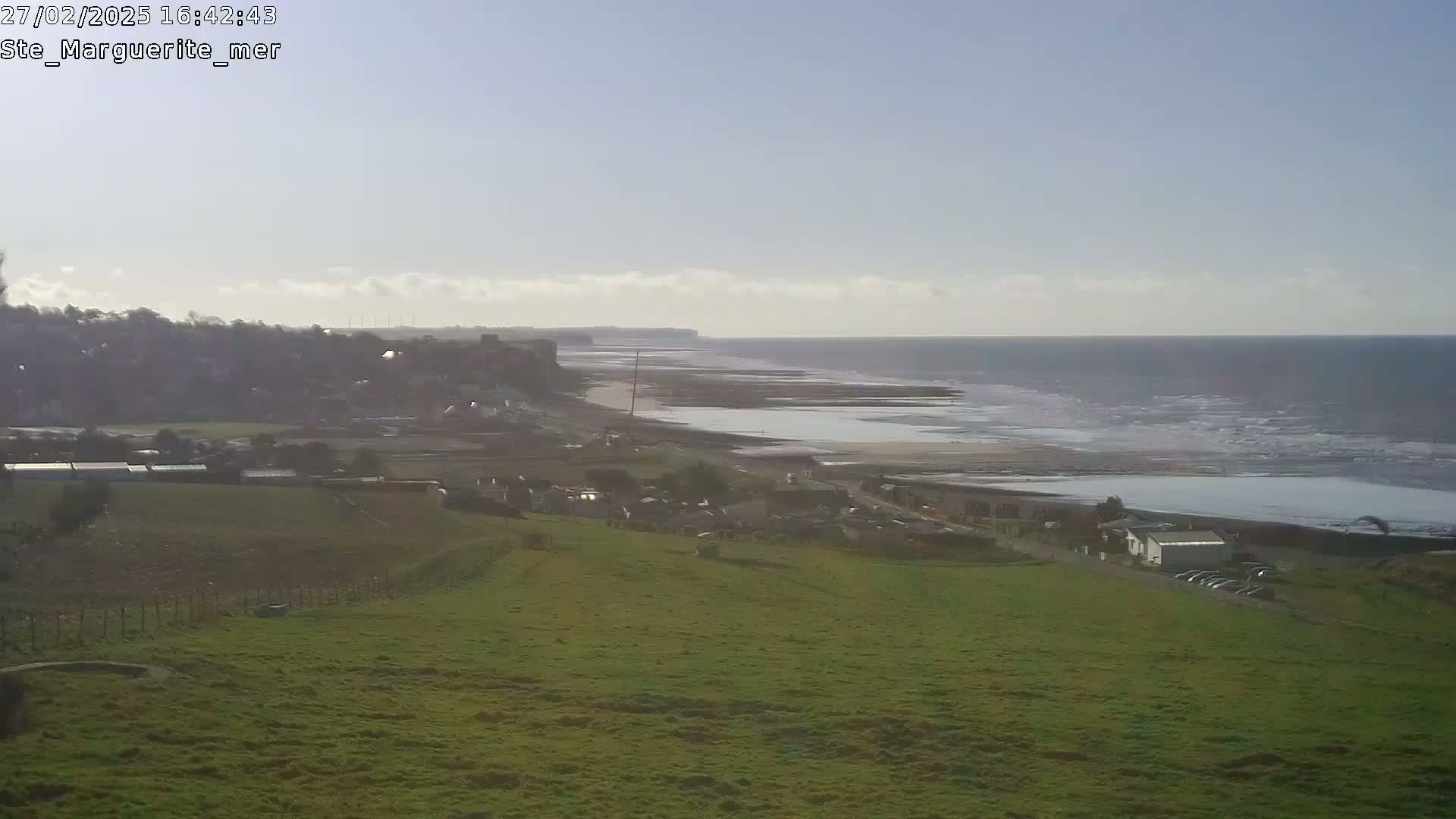 A coastal scene under a mostly clear sky shows a grassy hillside overlooking a village, a beach, and a calm sea with small waves.