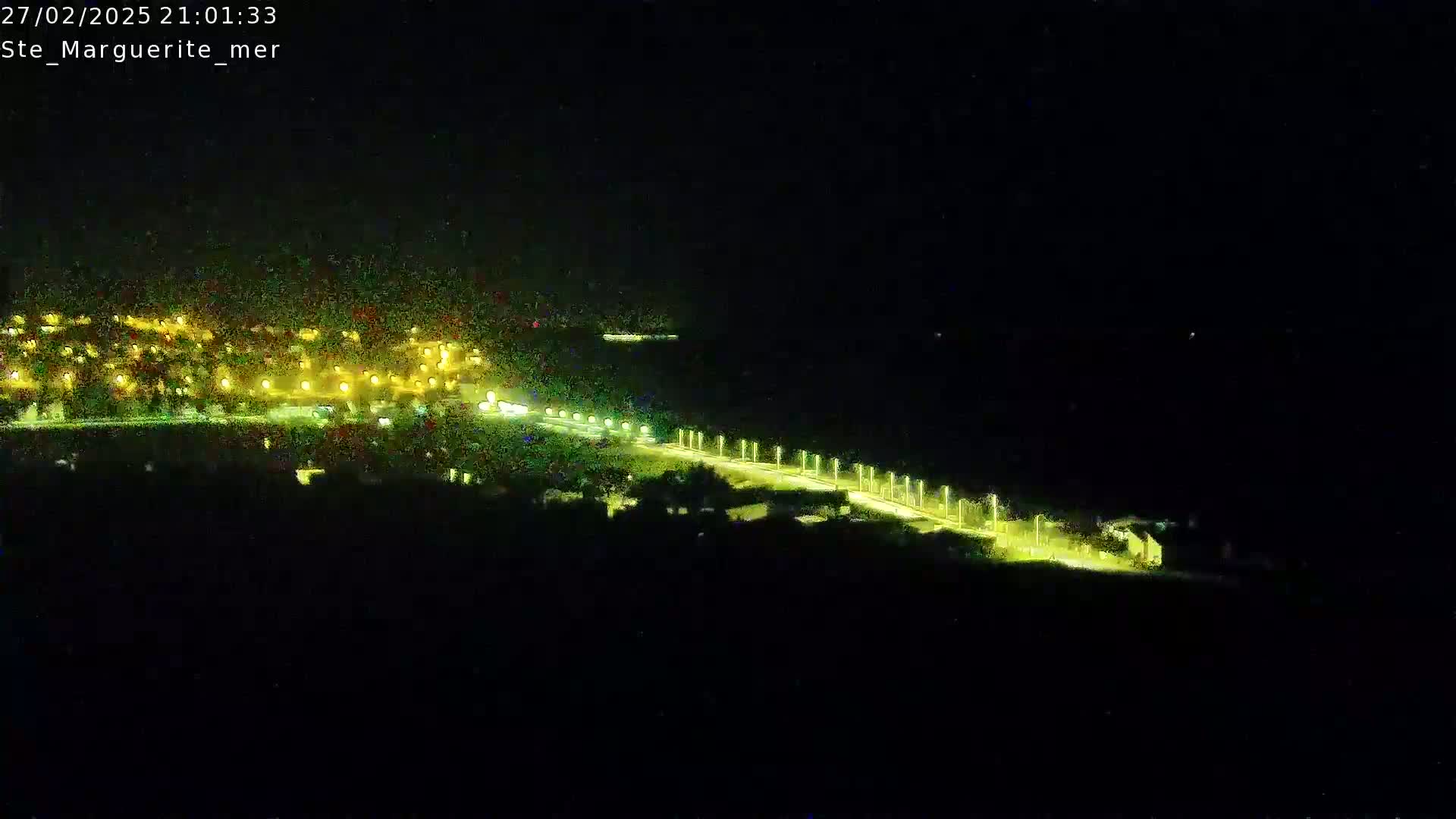 A nighttime aerial view shows a town's lights near a dark body of water, with a lit road curving along the water's edge.