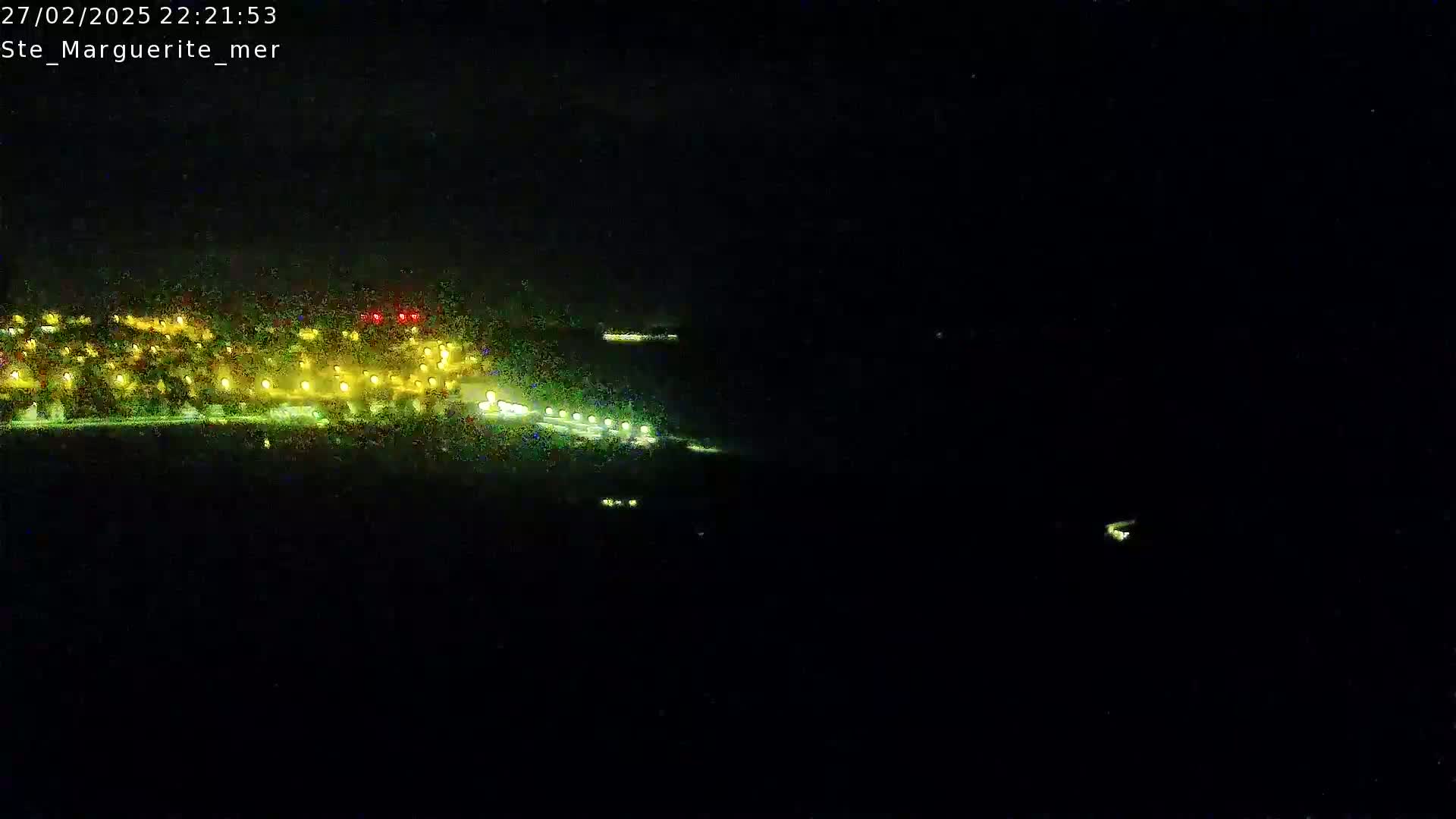 A nighttime long shot shows a coastal town with lights illuminating the buildings and a dark body of water in front, under a dark sky.