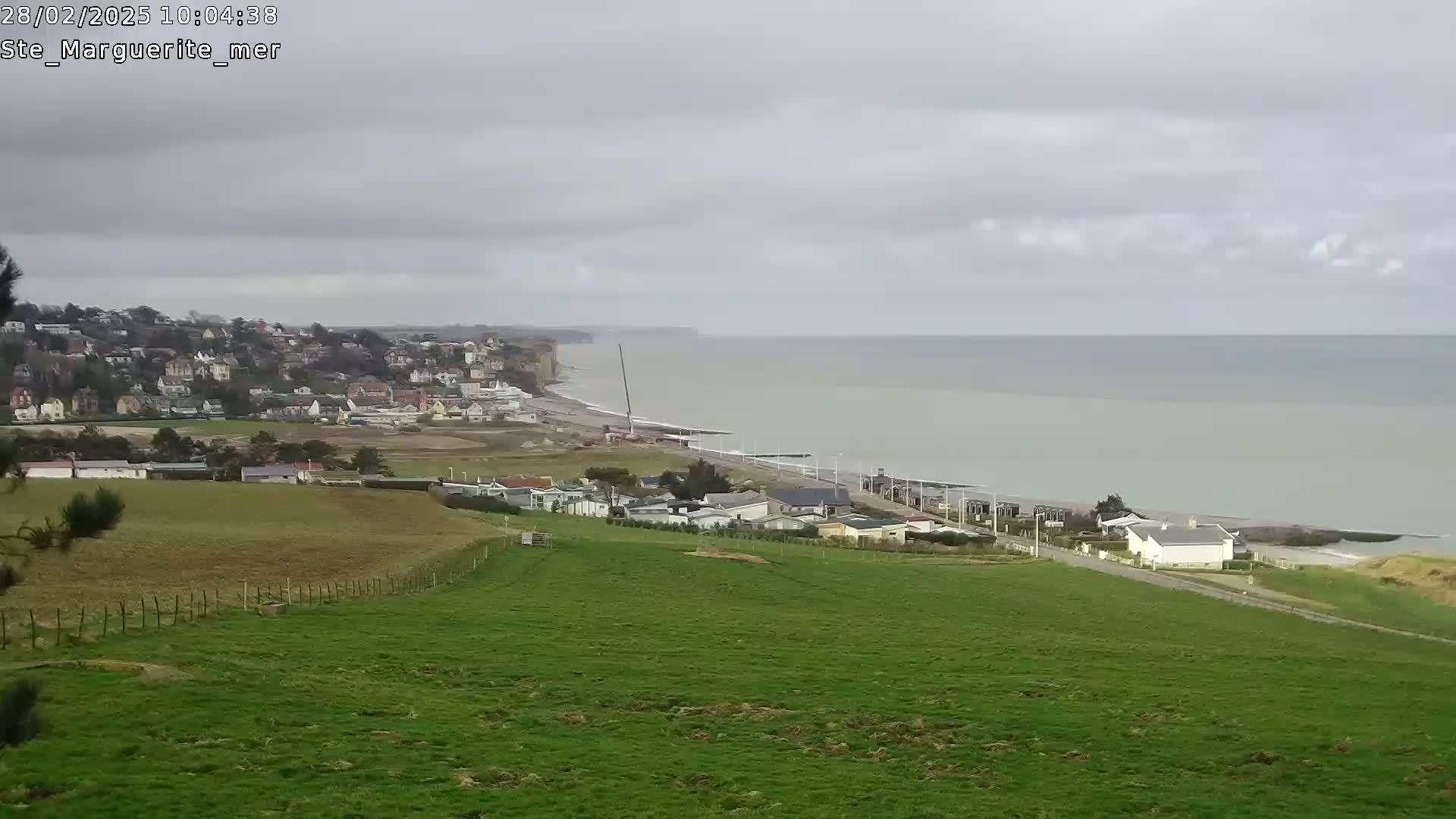 A coastal town sits on a hillside overlooking a calm sea under an overcast sky, with a green field in the foreground.
