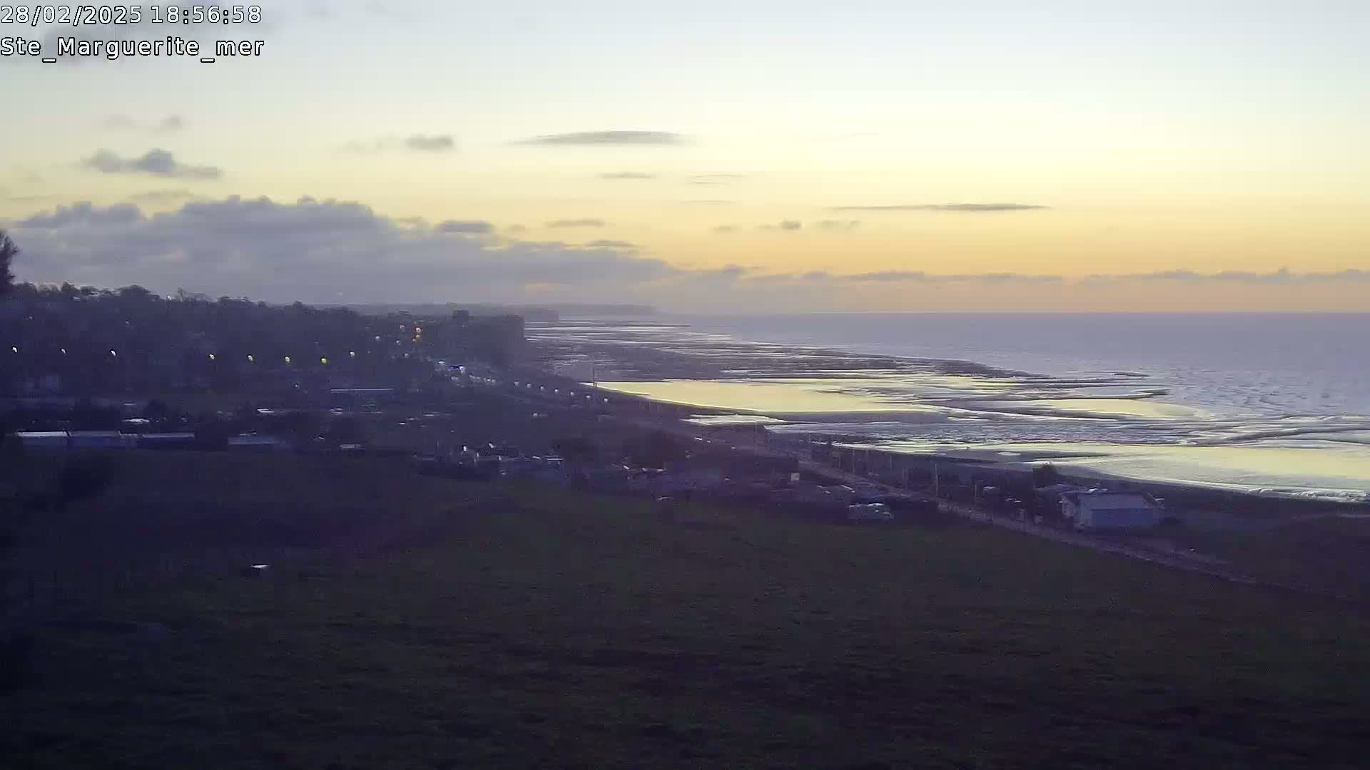 A coastal scene at sunset shows a town overlooking a beach and ocean with calm waves under a mostly clear sky.