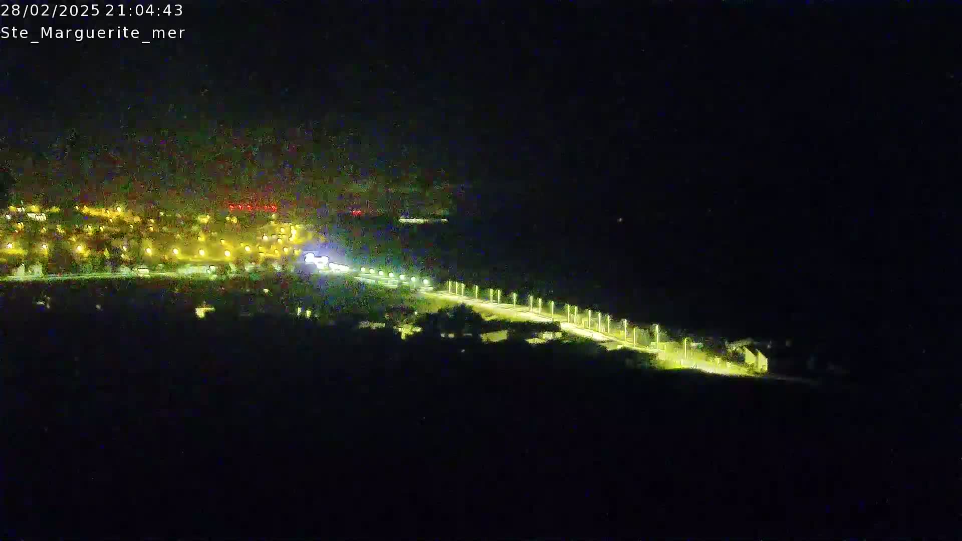 A nighttime aerial view shows a brightly lit road winding through a dark landscape toward a town illuminated with numerous lights.