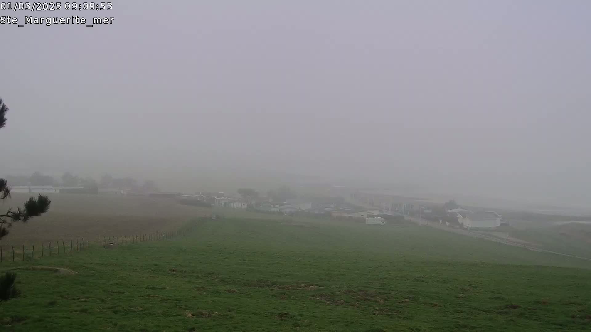 A foggy landscape reveals a distant village nestled beside a grassy field, viewed from a slightly elevated perspective.