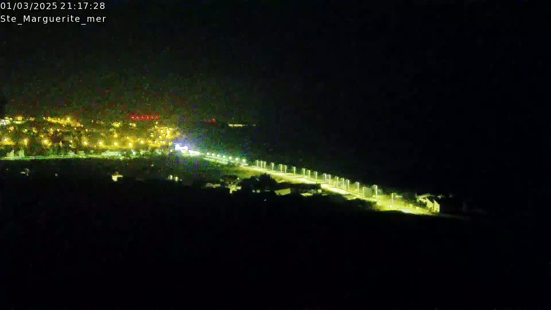 A nighttime, high-angle view shows a brightly lit road curving through a dark landscape towards a distant town illuminated by numerous lights.