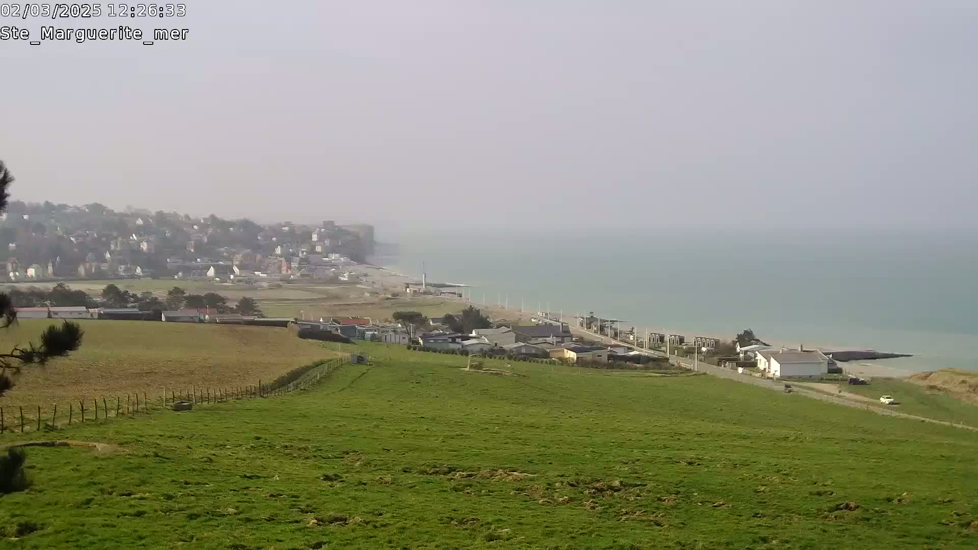 A hazy view shows a coastal town nestled below a grassy hill overlooking a calm sea.