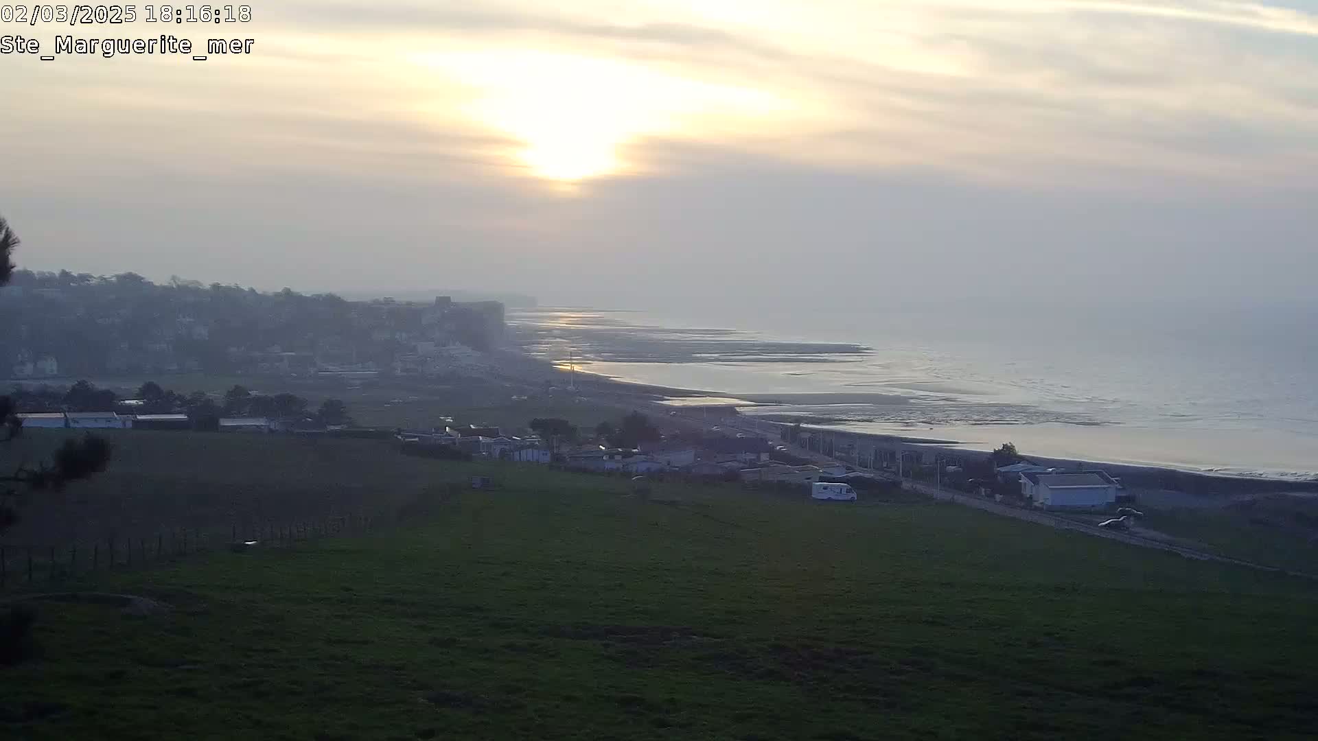 A hazy sunset over a coastline, showing a town nestled against hills overlooking a calm ocean with a sandy beach.