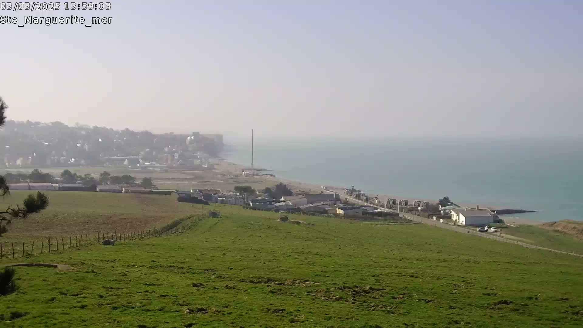 A hazy coastal scene shows a green hillside overlooking a town and a calm, teal sea.