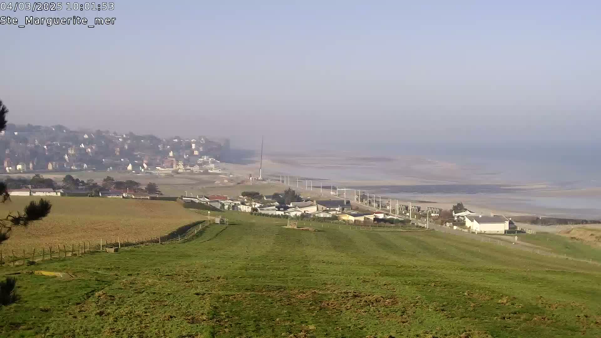 A hazy, distant view shows a coastal town, a beach, and a grassy hillside dotted with buildings under a clear sky.