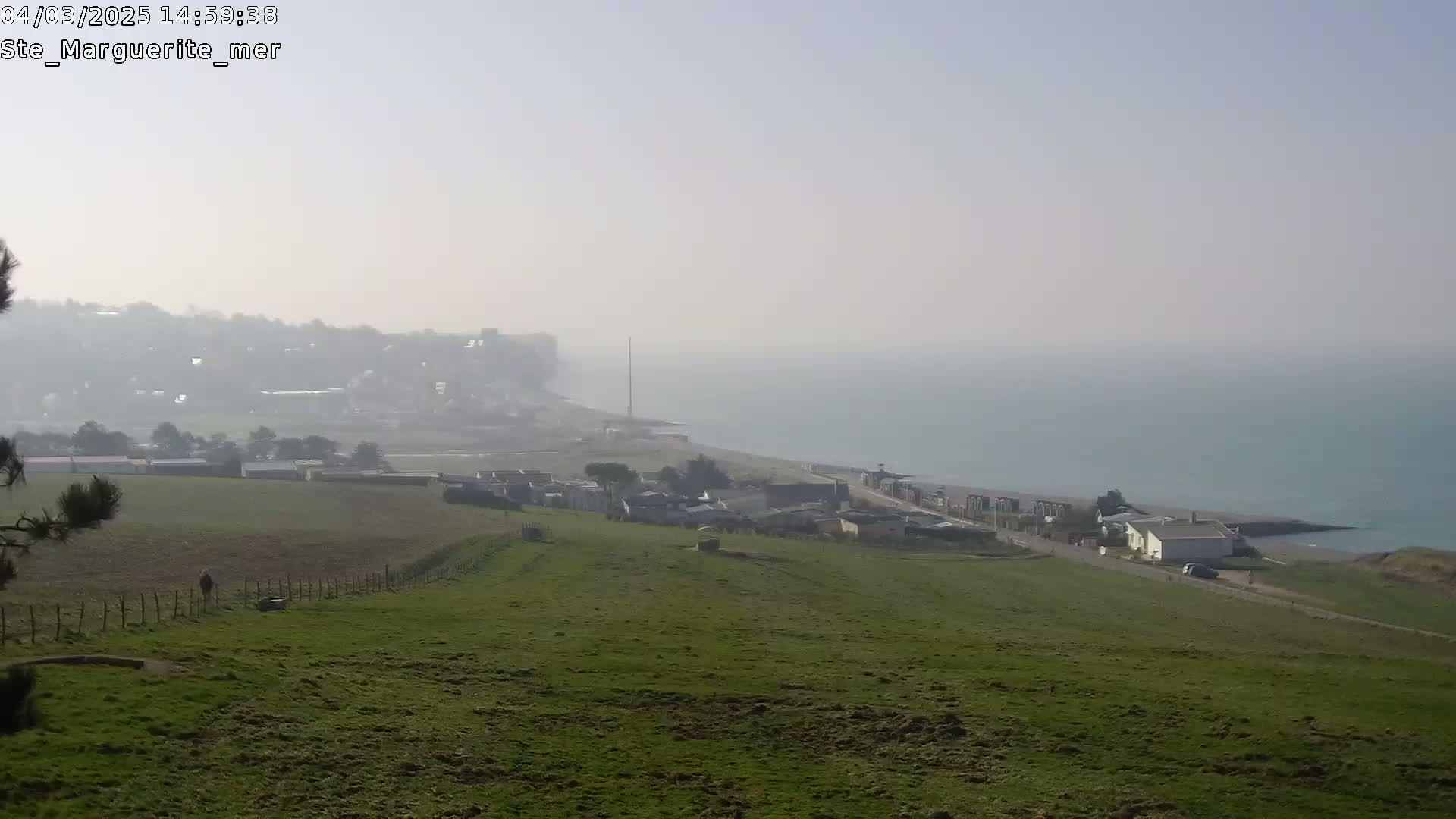 A hazy coastal scene shows a green hillside overlooking a village and ocean, with a person walking in a field in the foreground.