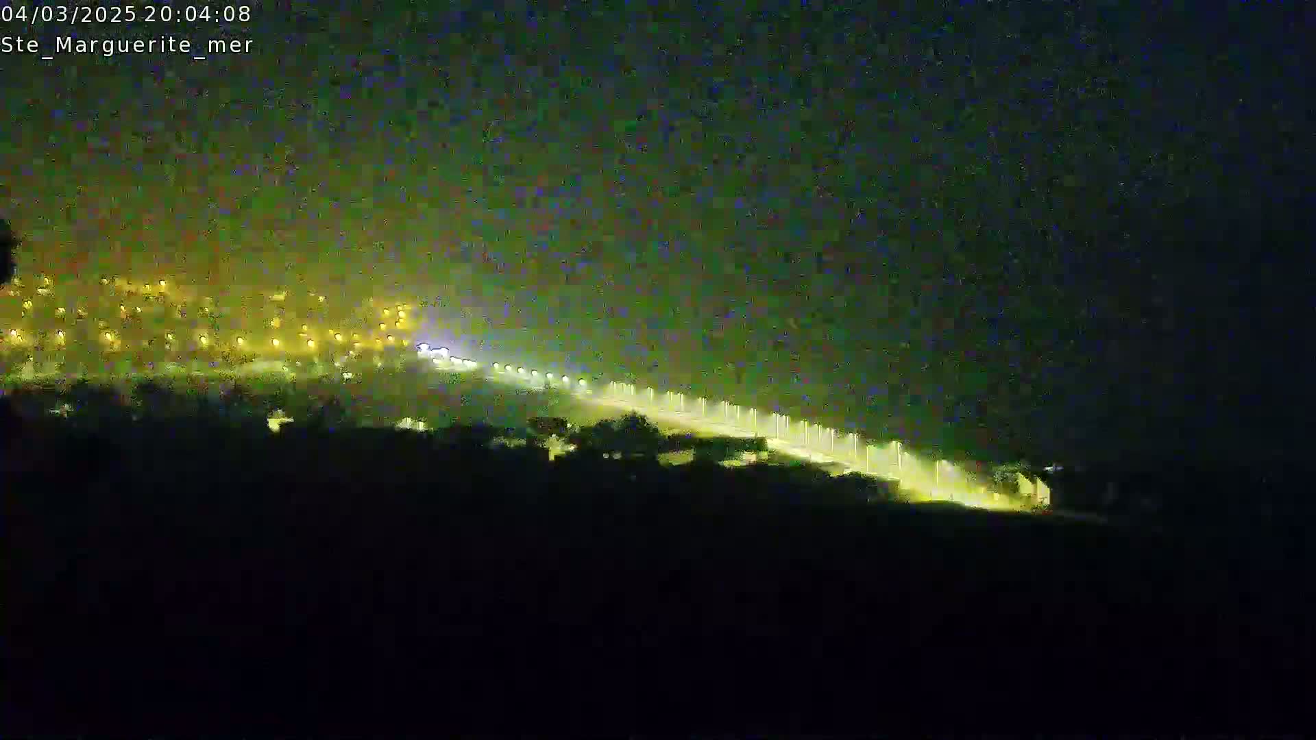 A nighttime long exposure shot shows a brightly lit road curving down a hill towards a town in the distance under a dark sky.