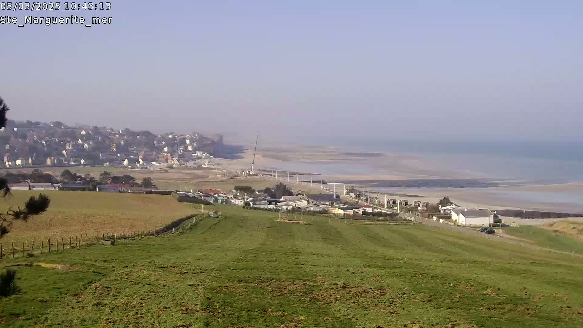 A grassy hillside overlooks a coastal town and a wide expanse of sandy beach and calm ocean under a clear sky.