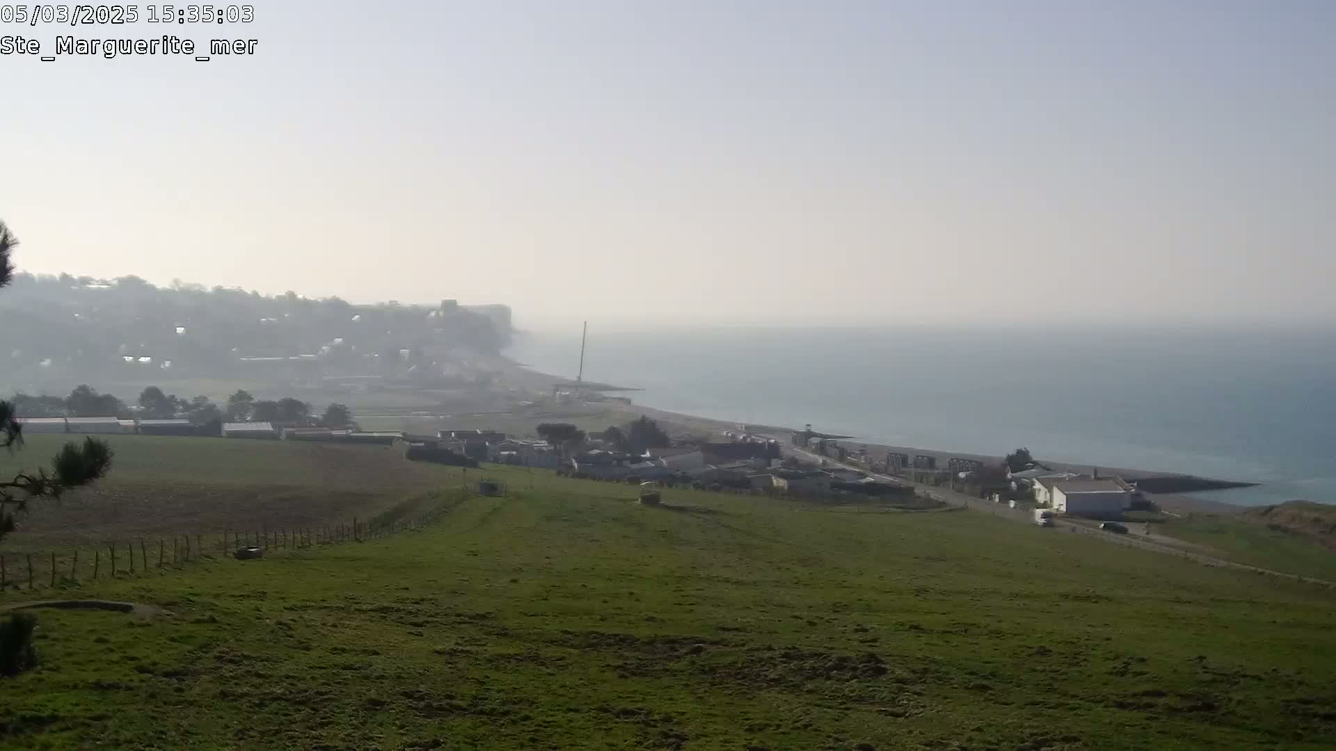 A hazy coastal view shows a green hillside overlooking a town, a beach, and a calm sea under a pale sky.