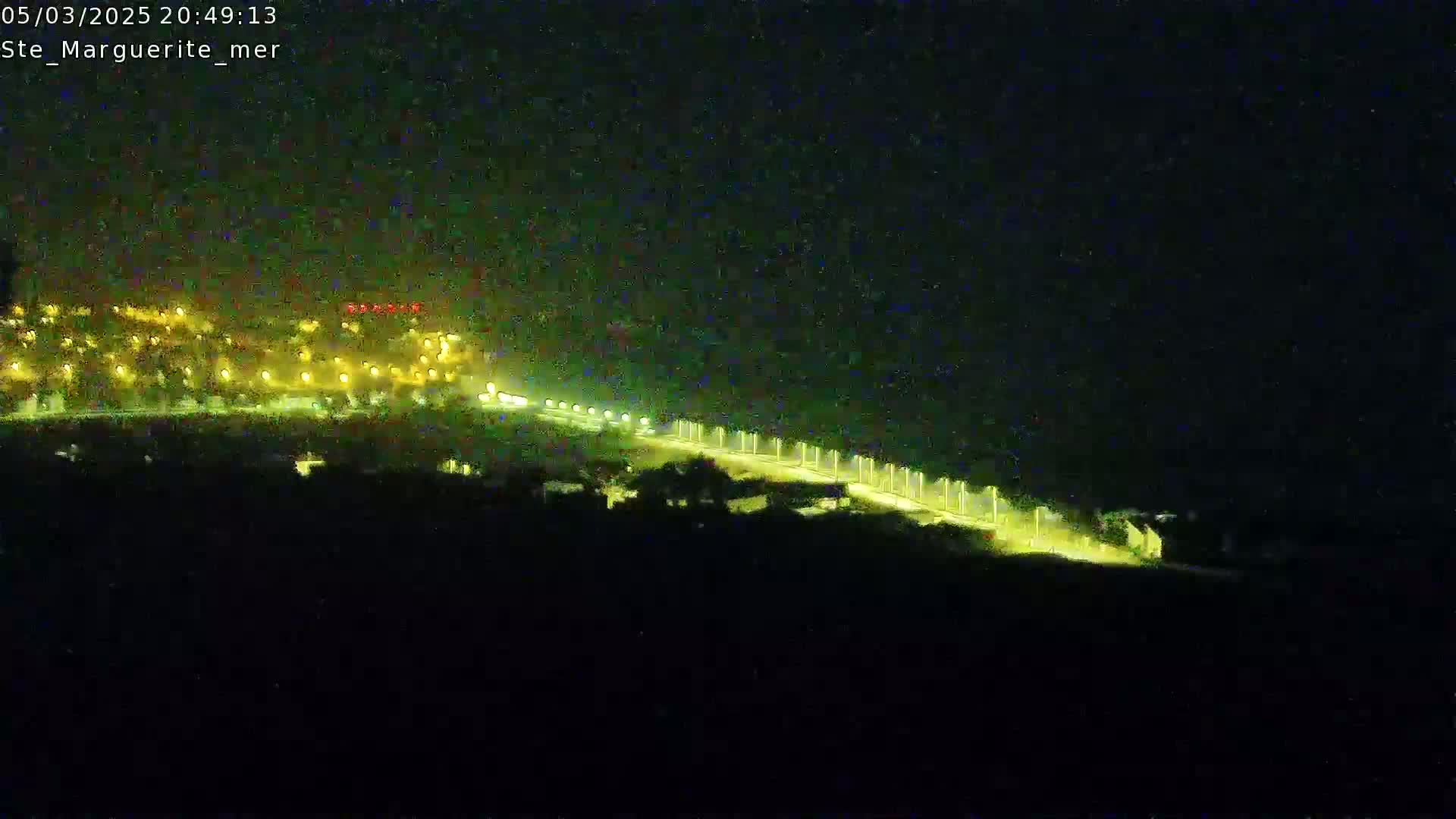 A nighttime, long shot shows a brightly lit road winding through a dark landscape with a town illuminated in the distance.