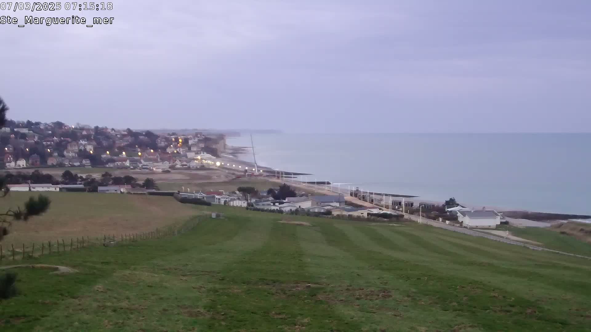 A coastal town sits below a grassy hillside overlooking a calm sea under an overcast sky.