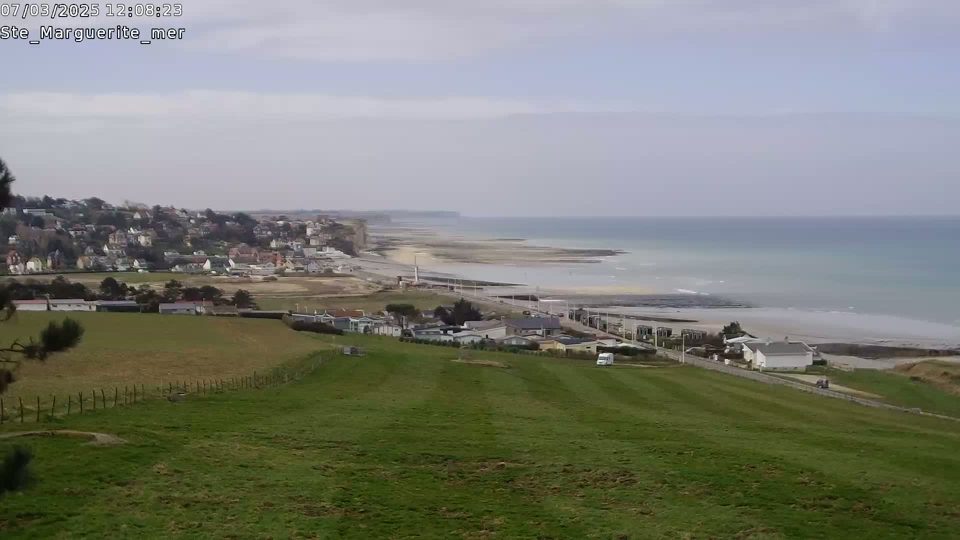 A coastal town nestled against high cliffs overlooks a long, sandy beach and calm ocean under an overcast sky.