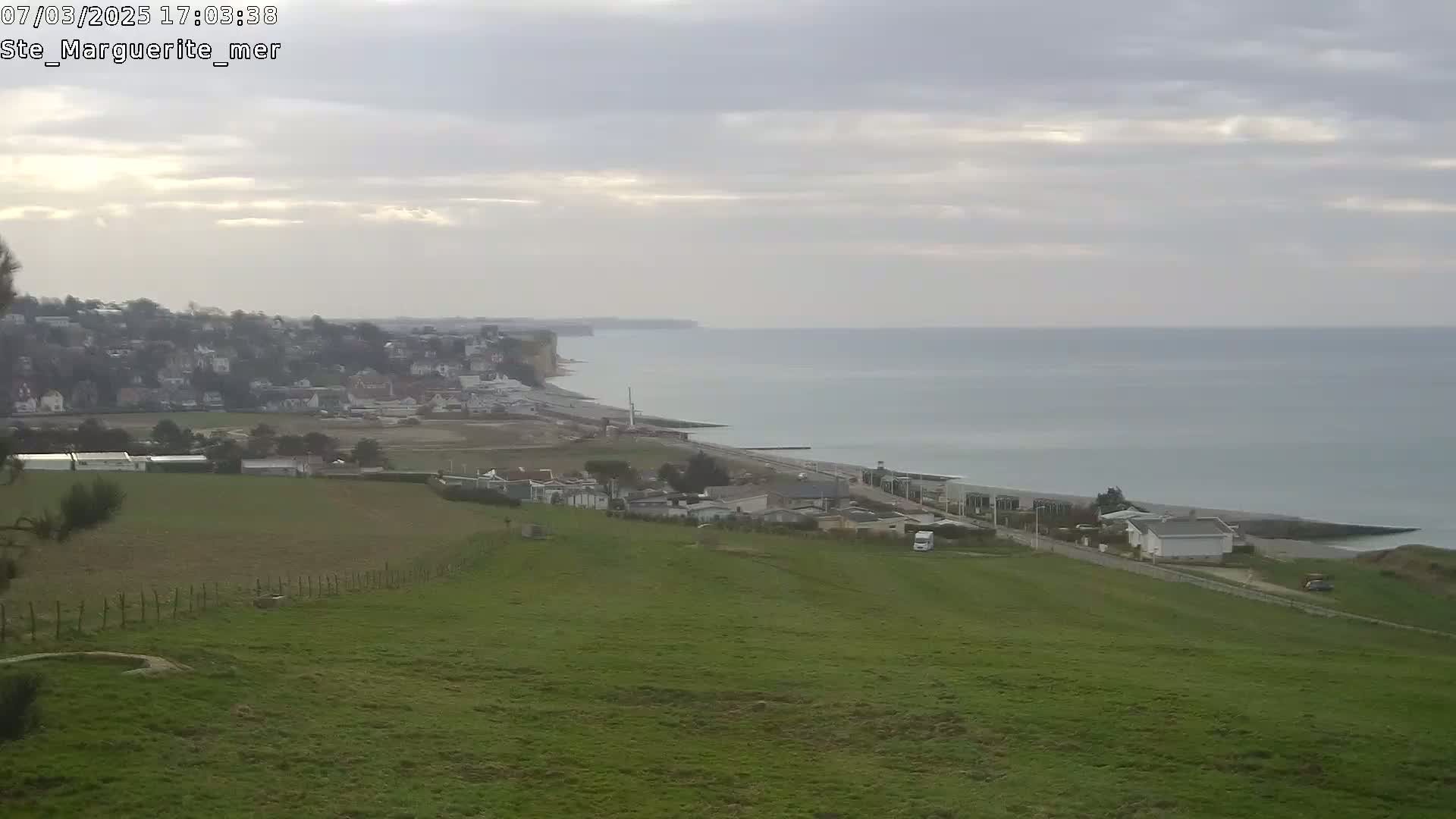 A coastal town sits below a grassy hill overlooking a calm sea under an overcast sky.