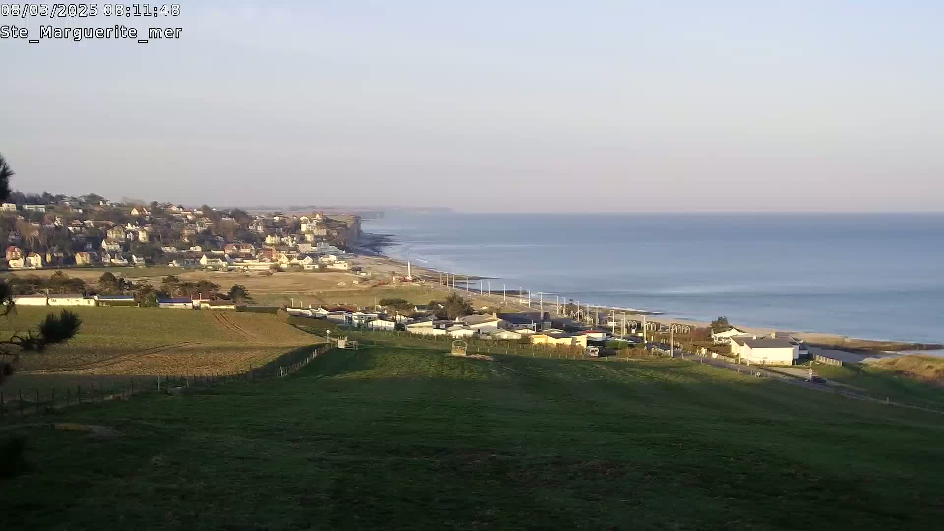 A coastal town with houses nestled on a hillside overlooks a calm ocean under a clear sky.