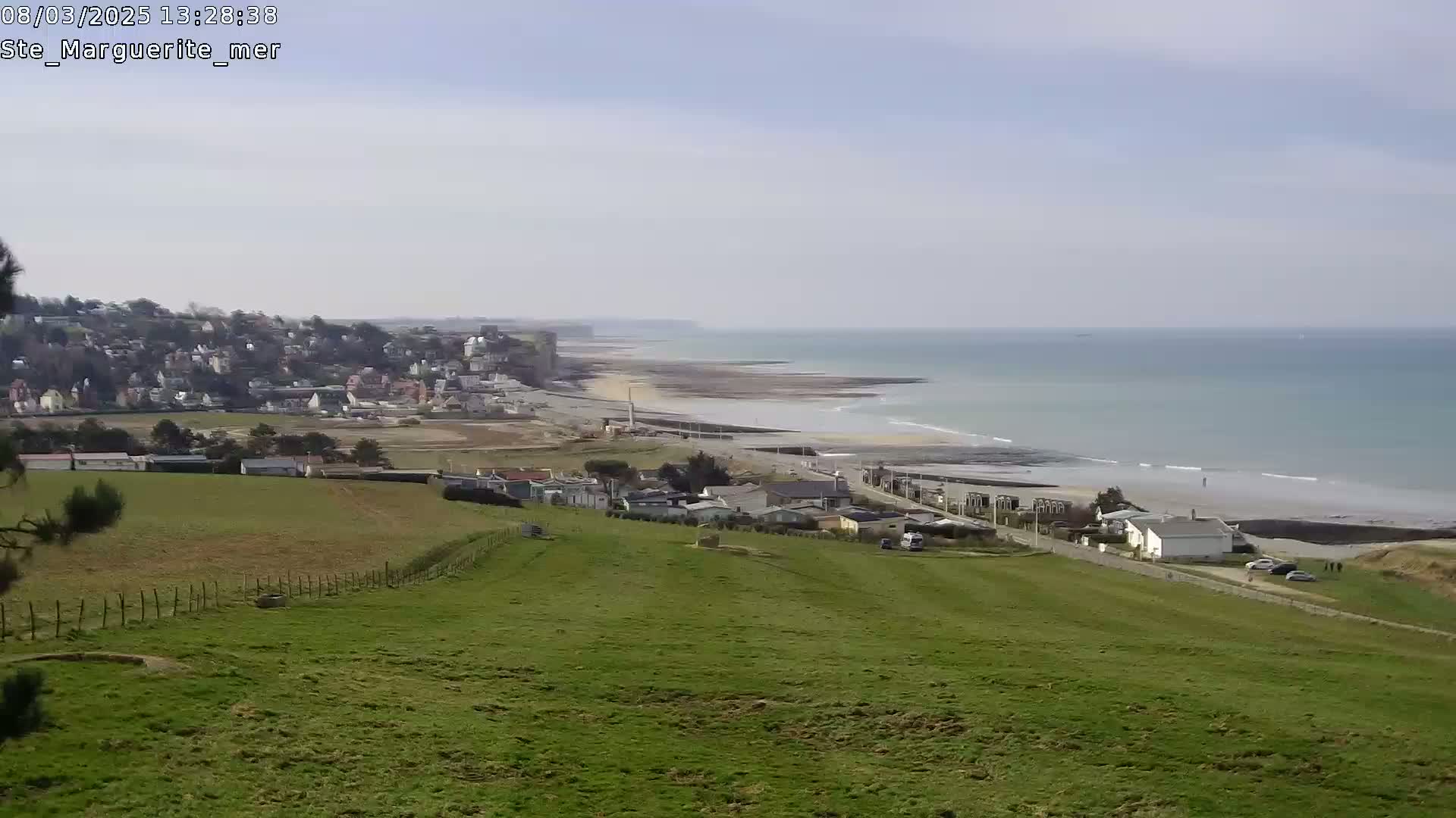 A coastal town sits on a hillside overlooking a long, sandy beach and calm ocean under a mostly clear sky.