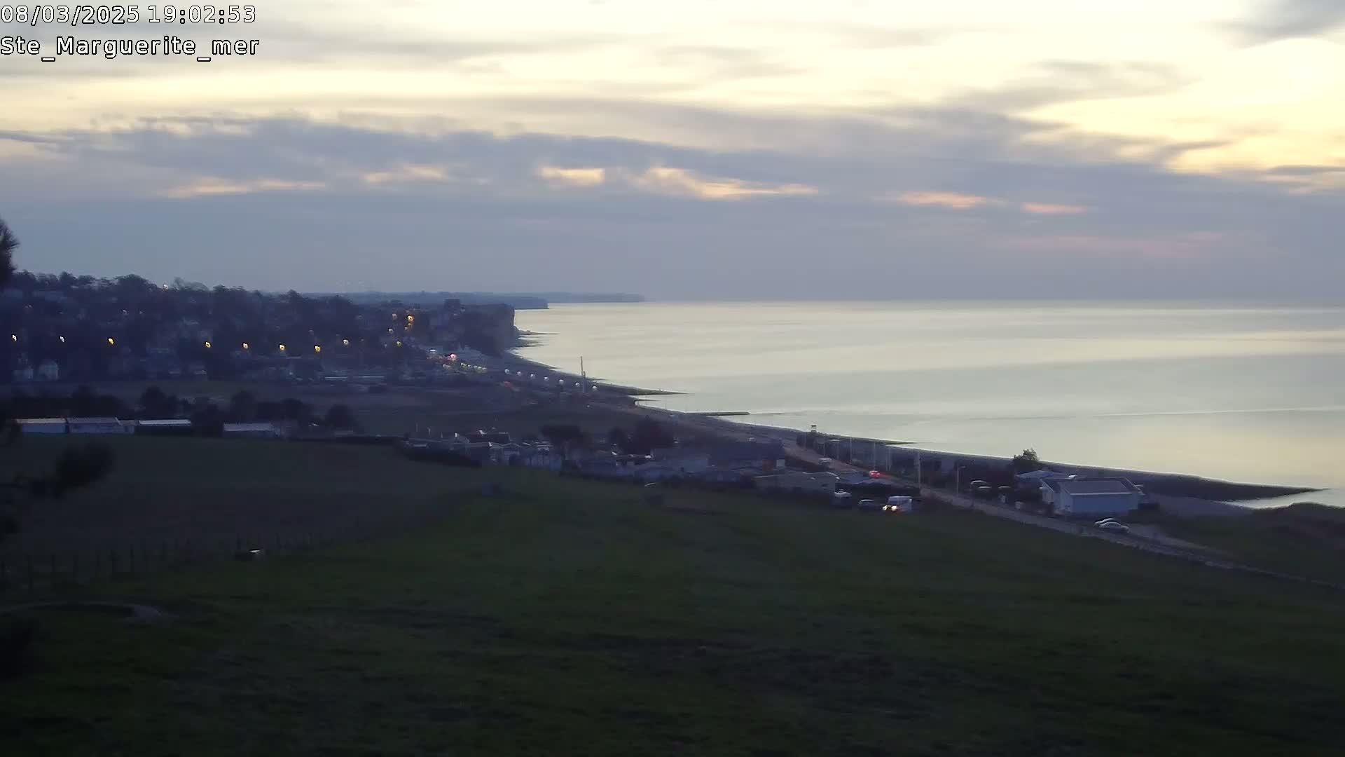 A coastal town at dusk is visible from a hilltop, overlooking a calm sea under a mostly cloudy sky.