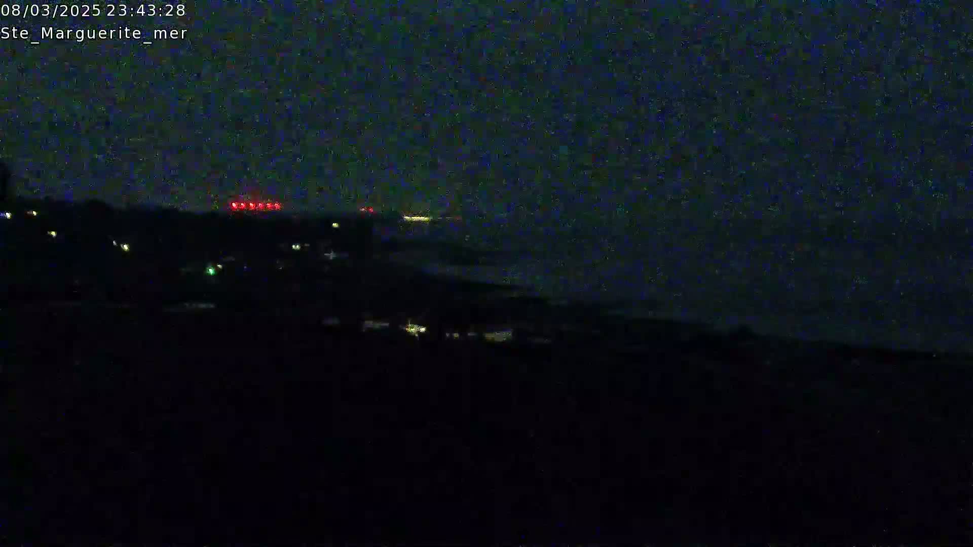 A nighttime, dark coastal scene shows a faint horizon line, a few scattered lights on land, and a line of red lights possibly from a distant ship or structure on the water.