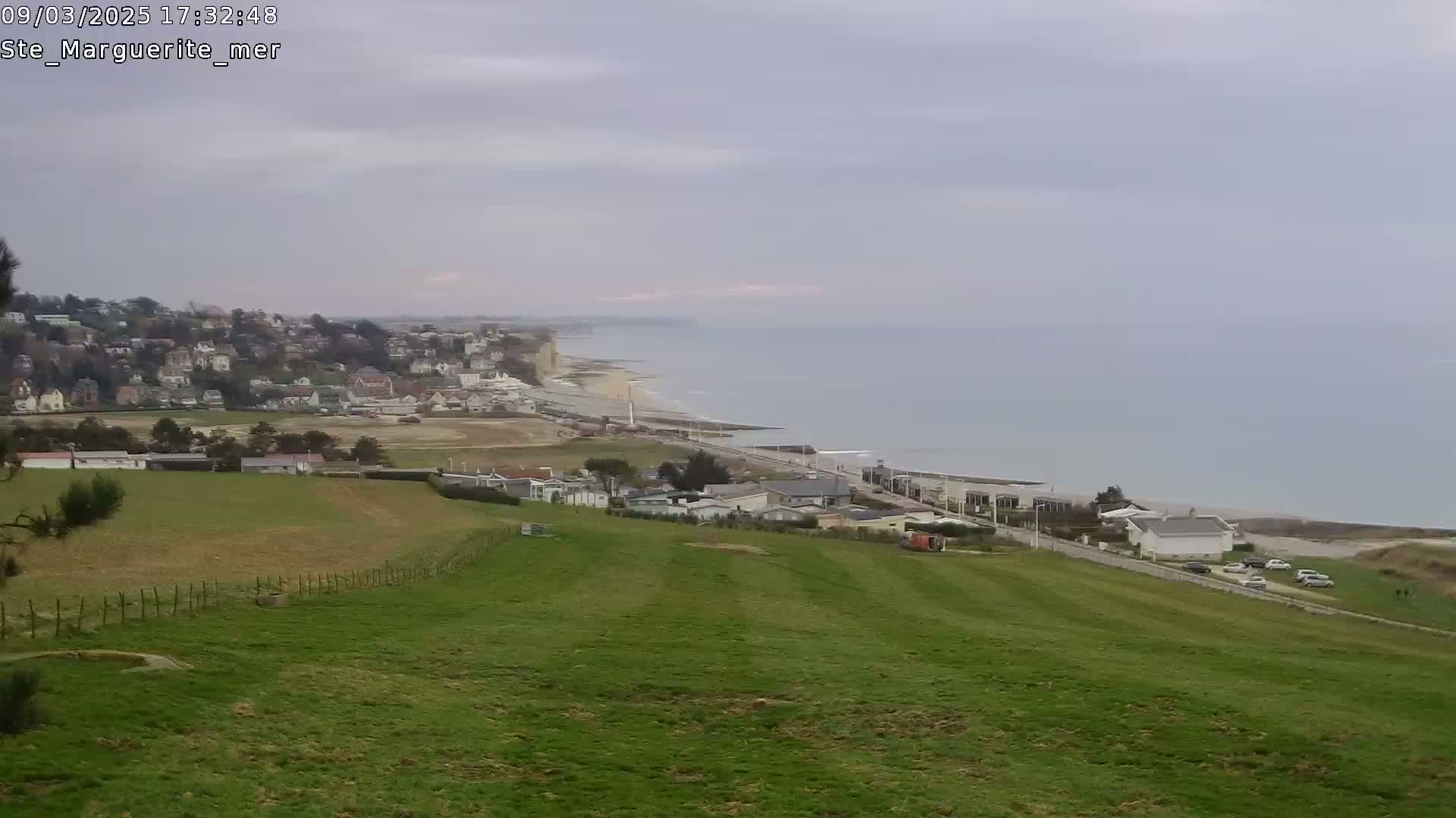 A coastal town and beach are visible from a grassy hillside under an overcast sky.
