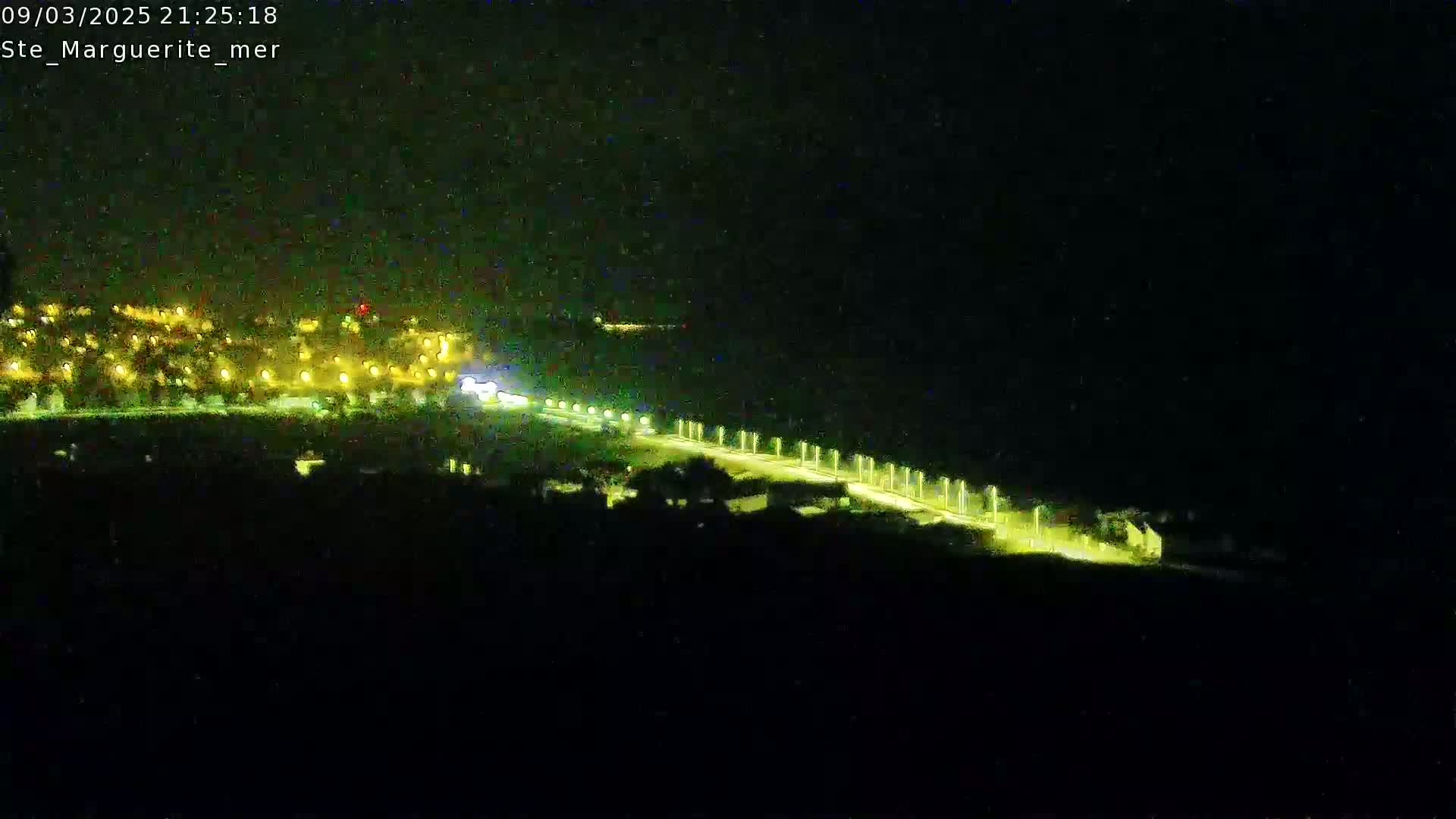 A nighttime, long shot shows a coastal road with illuminated lights curving away from a town whose lights are visible in the distance.