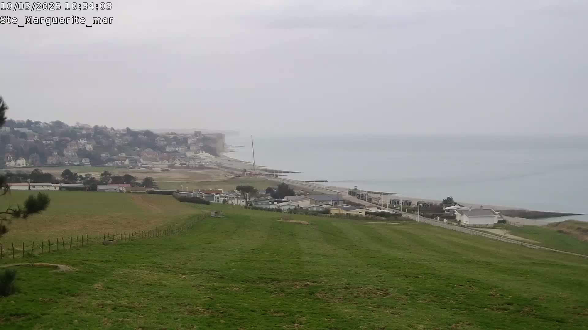 A coastal town sits below a grassy hill overlooking a hazy, grey ocean under an overcast sky.