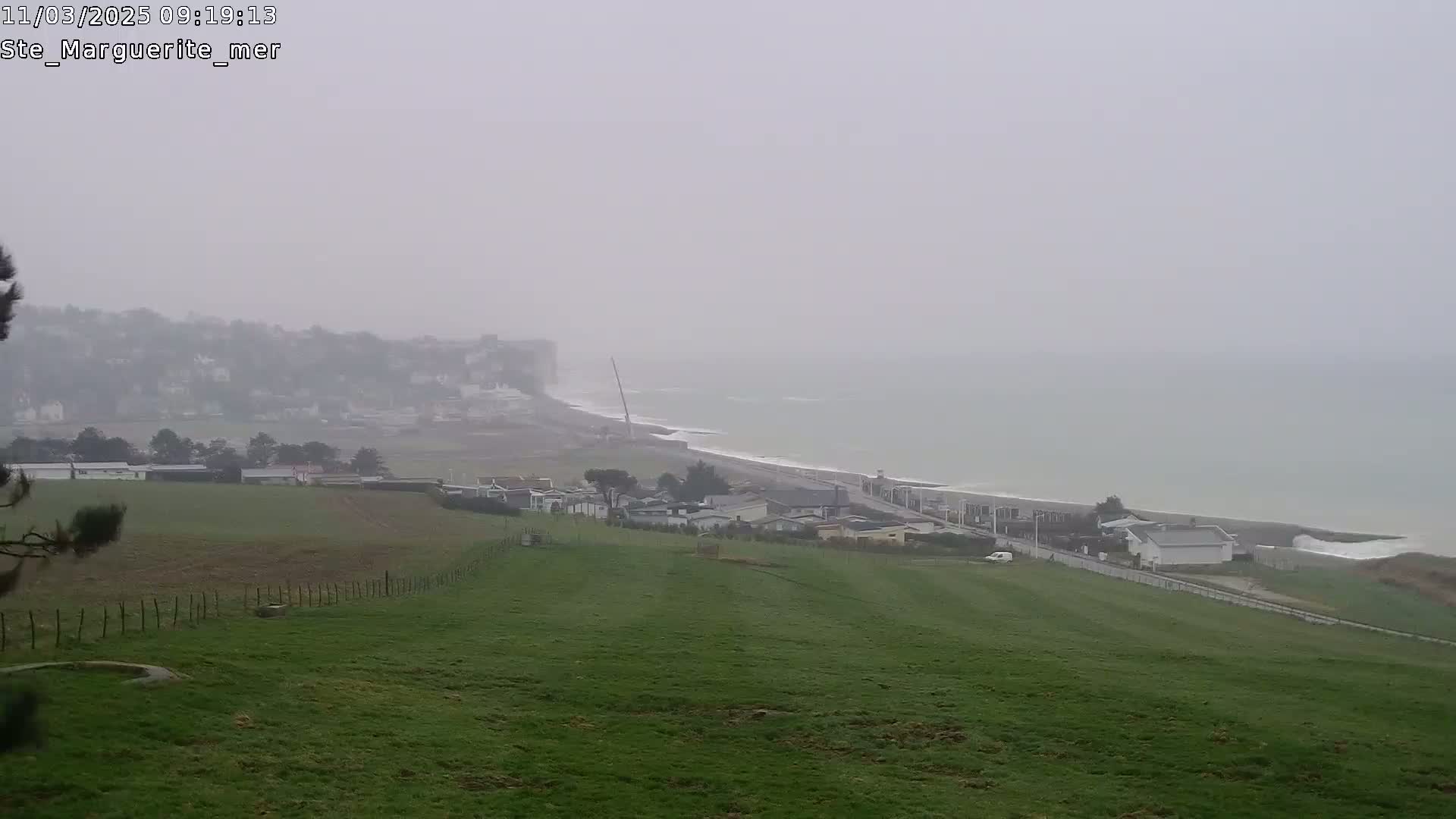 A foggy coastal scene shows a green field sloping down to a road, houses, a beach, and a hazy ocean.
