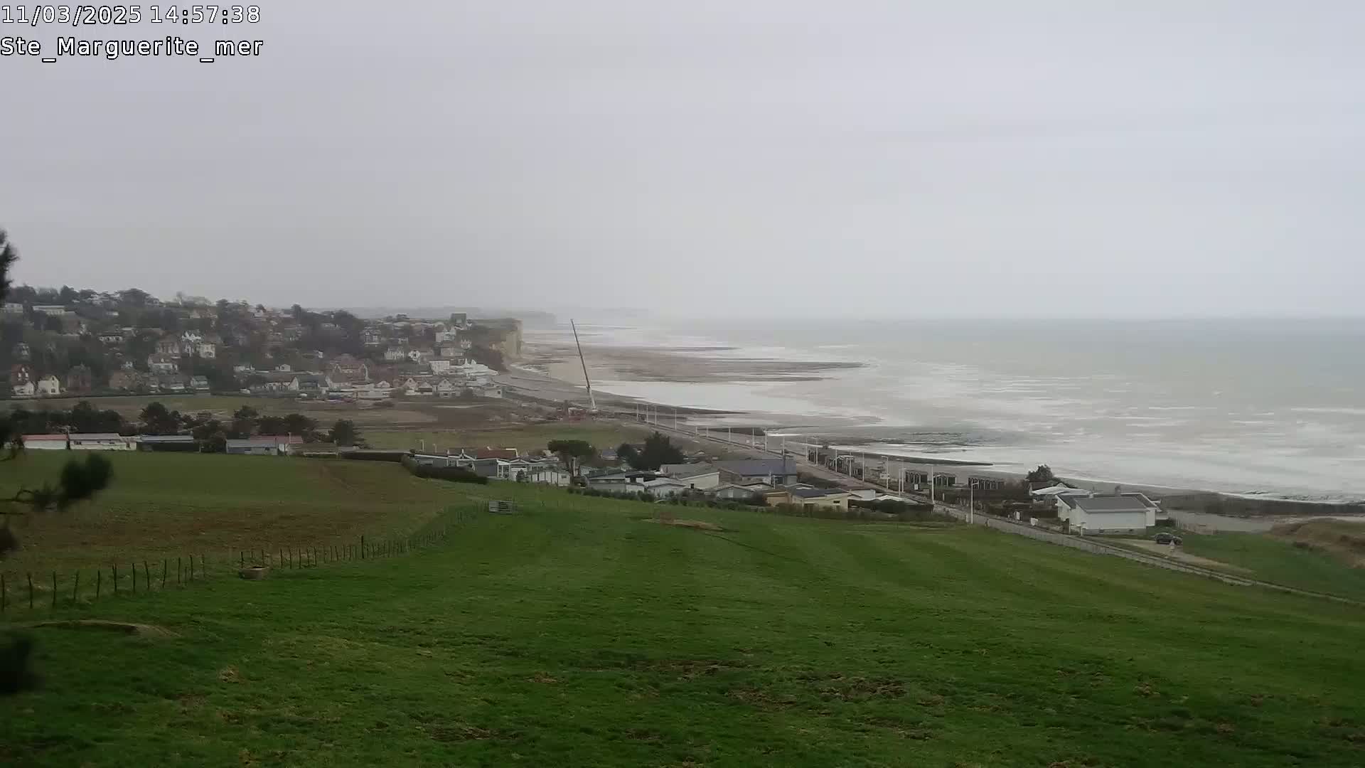 A grassy hillside overlooks a coastal town and a grey, overcast ocean with waves breaking on the shore.