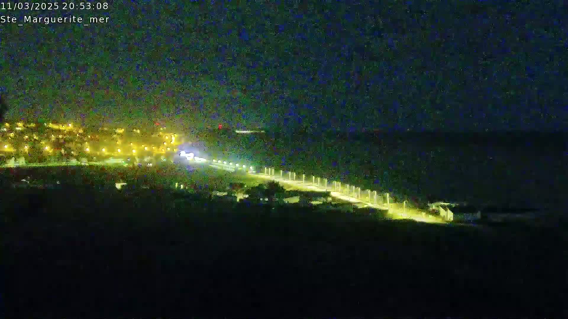 A nighttime, long-range view shows a brightly lit road curving along a dark body of water, with a town's lights visible in the distance.