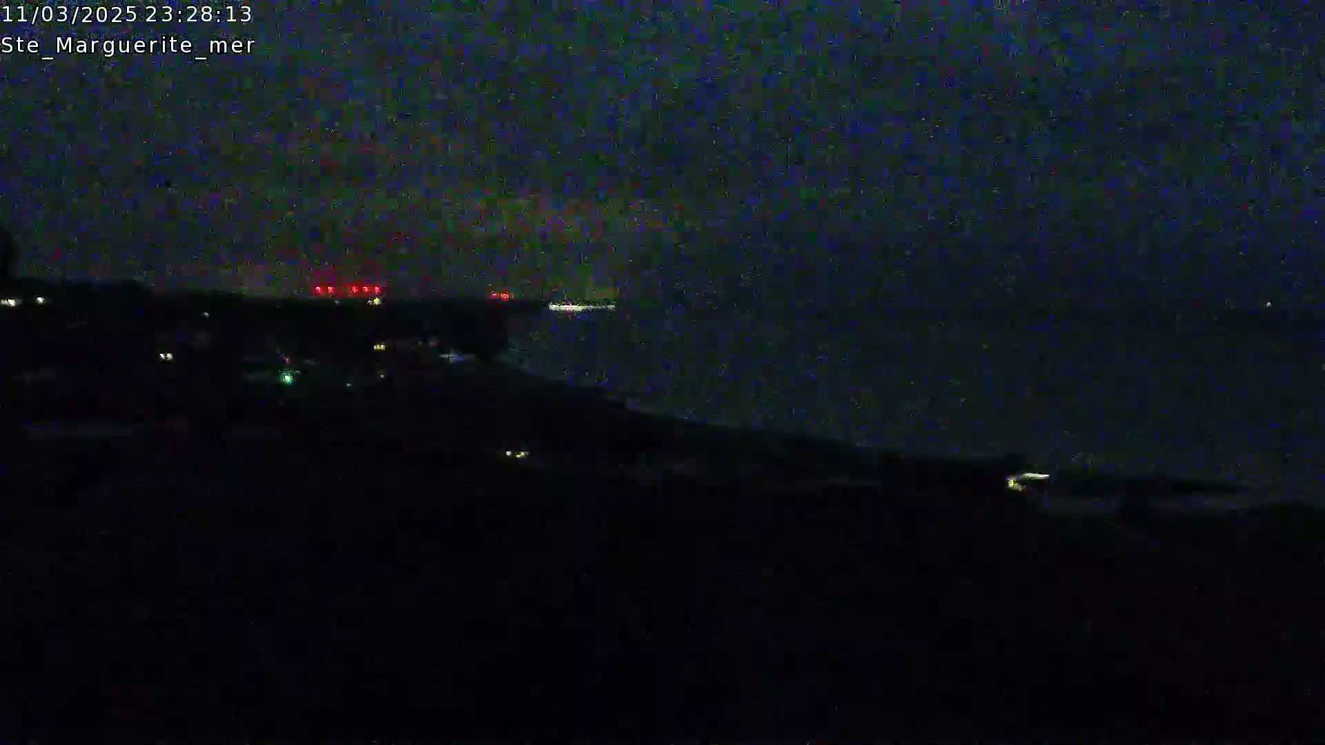 A nighttime, dark image shows a body of water with a few distant lights on the horizon and a few faint lights closer to the foreground on land.