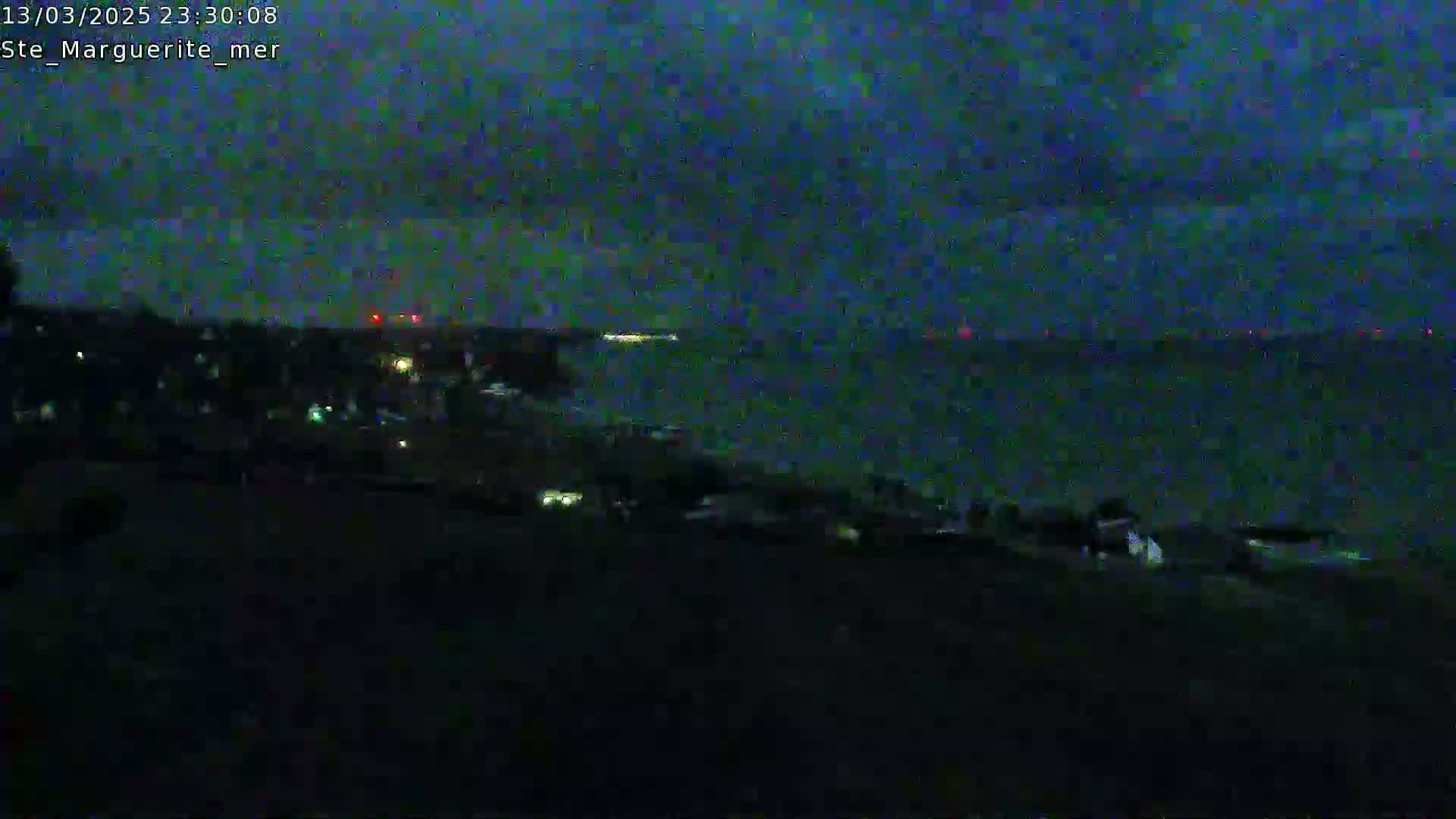 A dark, nighttime view shows a body of water under a mostly clear, dark sky, with a shoreline in the foreground featuring dimly lit buildings and a few lights on the beach.