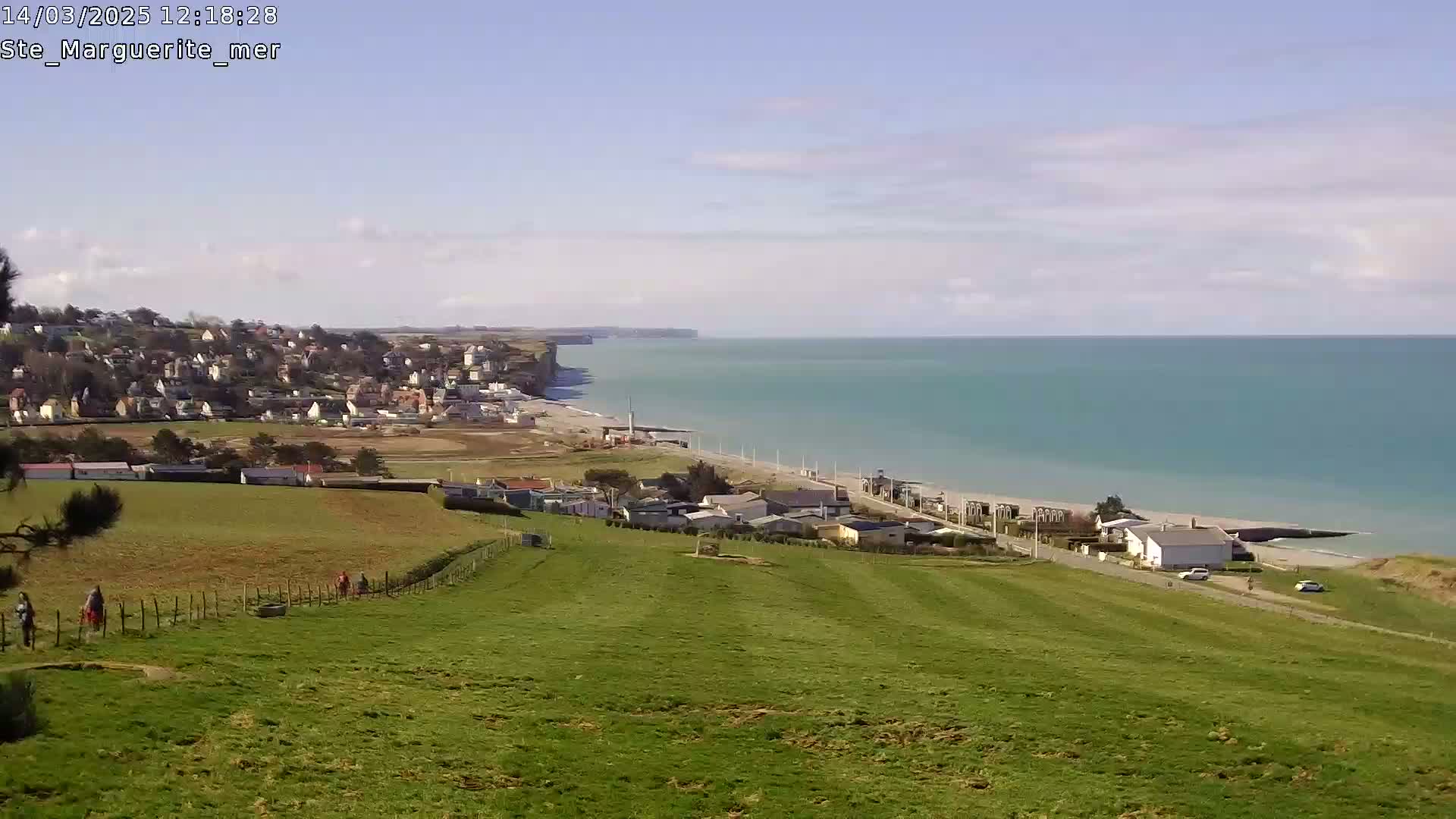 A coastal town sits atop cliffs overlooking a calm, teal ocean under a mostly clear, bright sky; a grassy hillside slopes gently down from the foreground towards the shore.