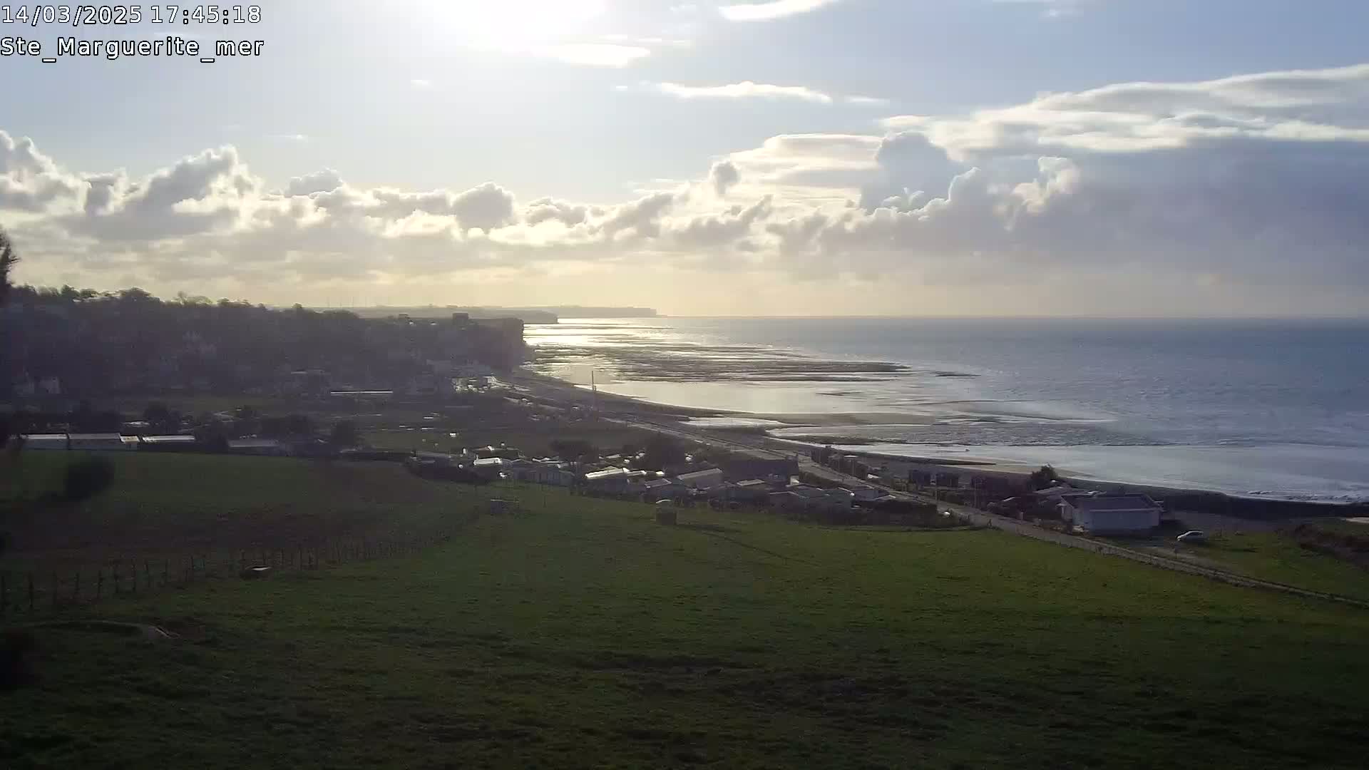 A coastal scene under a partly cloudy sky shows a town nestled below a grassy hill overlooking a beach and ocean with a cliff visible in the distance.