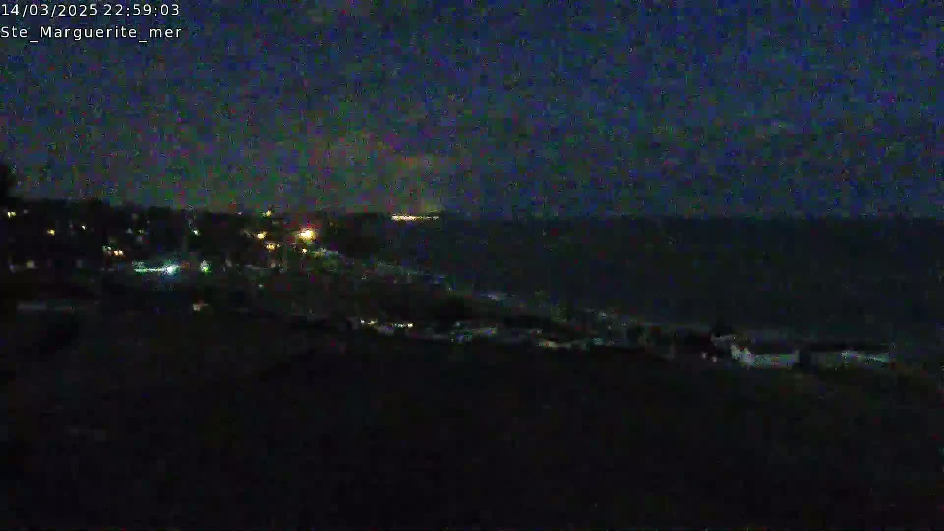 A dark, nighttime view shows a coastline with a few lights visible in the distance, overlooking a dark ocean under a mostly clear sky.