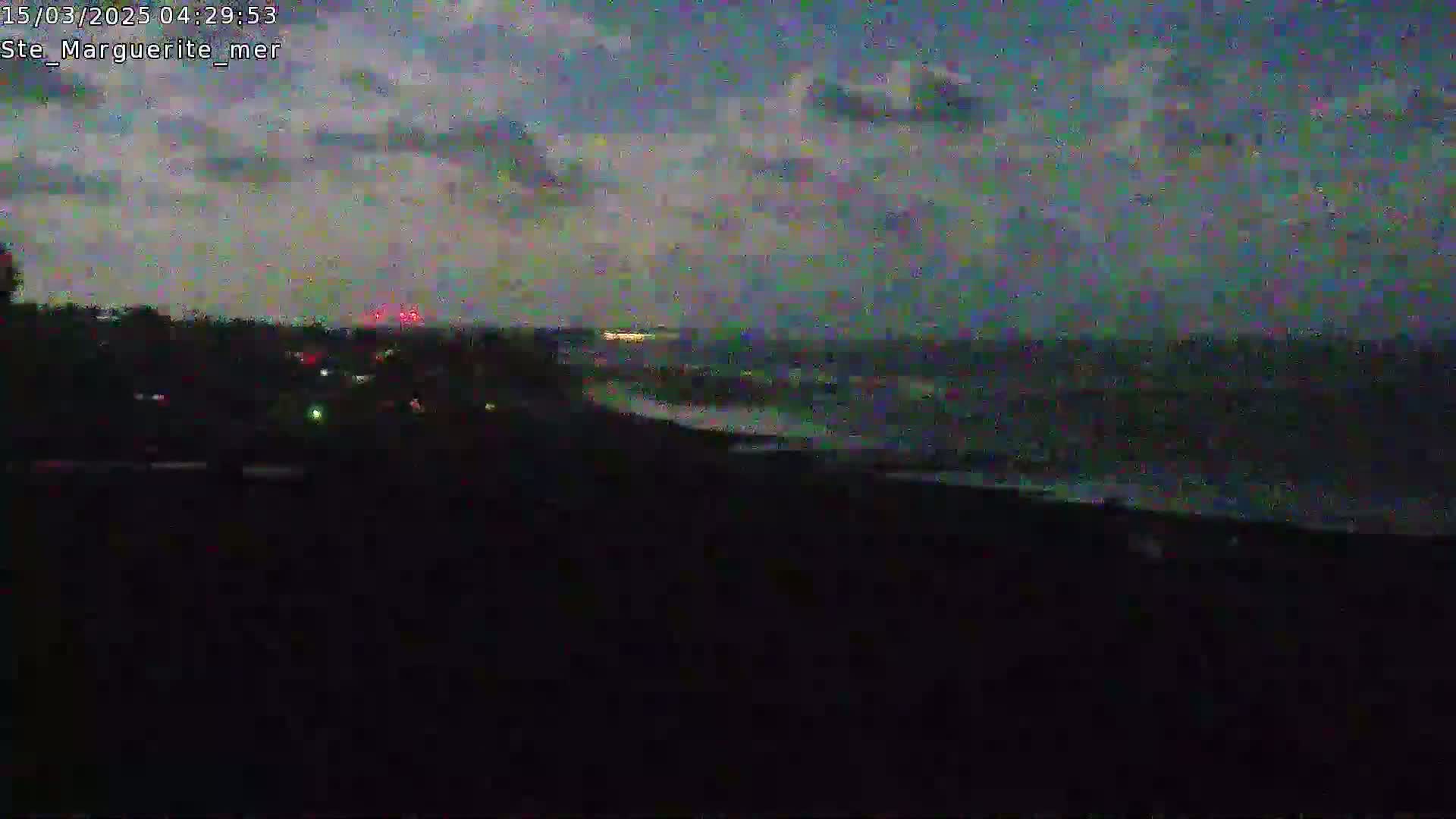 A dark, nighttime view shows a wavy ocean under a cloudy sky, with a dark shoreline and faintly lit land visible in the distance.