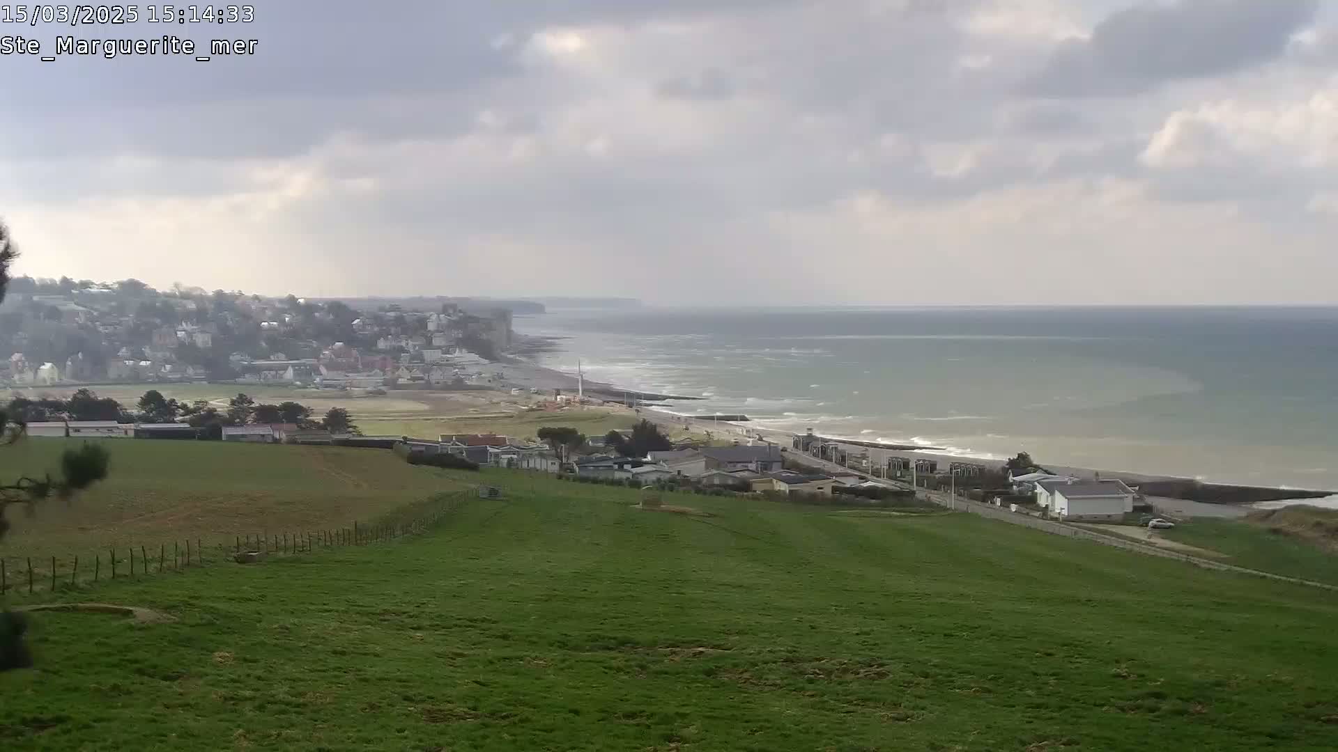 A sprawling green field overlooks a coastal town and a choppy sea under a cloudy sky.