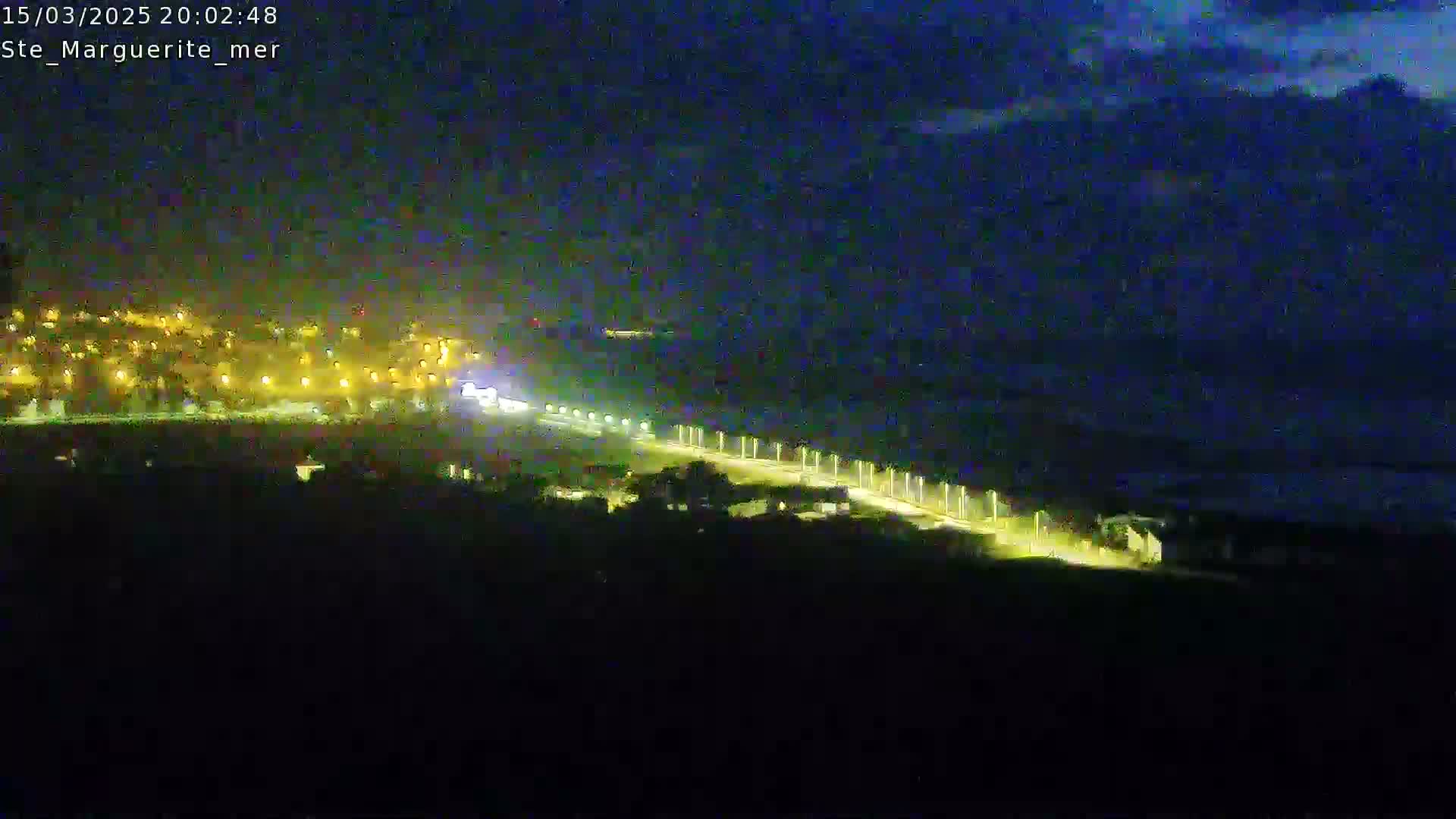 A nighttime, high-angle view shows a coastline with a town illuminated by streetlights extending along a road beside a dark body of water under a mostly clear night sky.