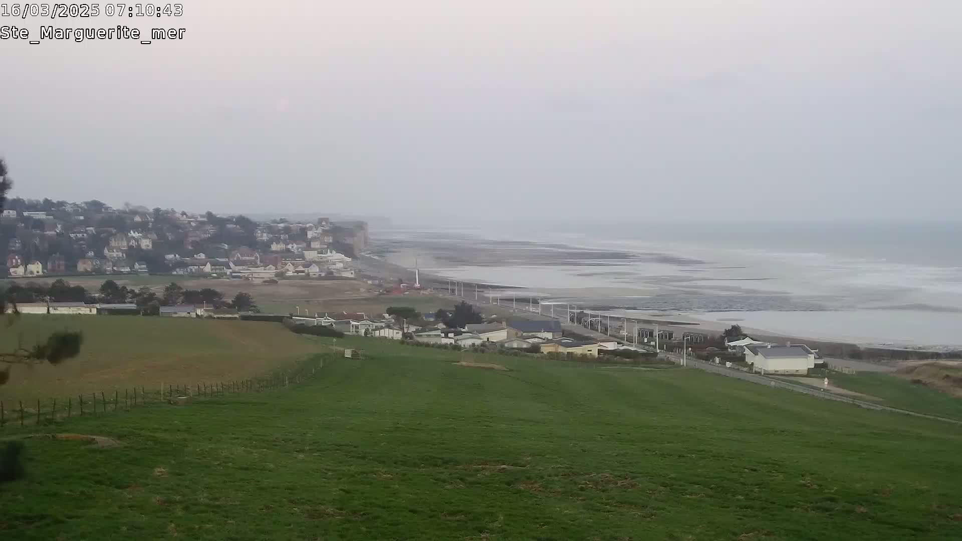 A hazy, overcast day reveals a coastal town nestled beside a wide, sandy beach that stretches to a calm sea under a pale sky.