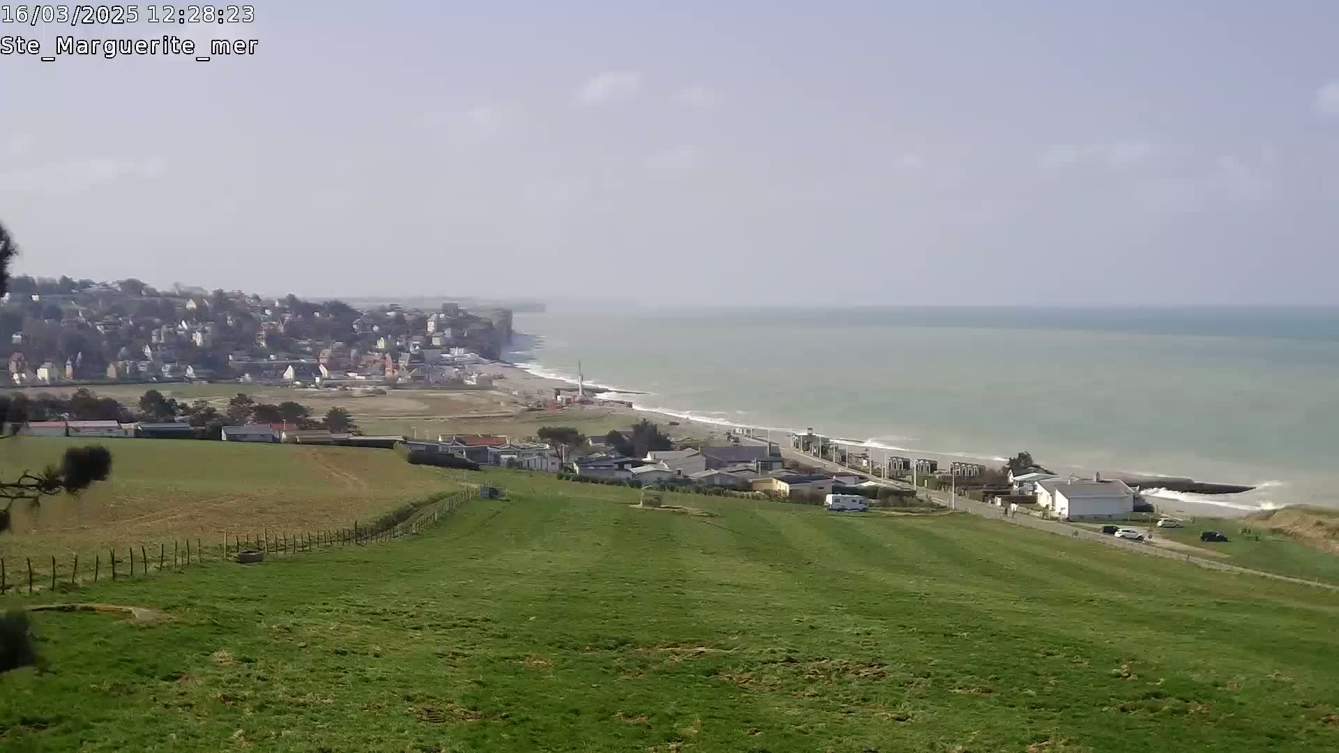 A coastal town nestled against a cliff overlooking a calm, light-blue ocean under a mostly clear sky.